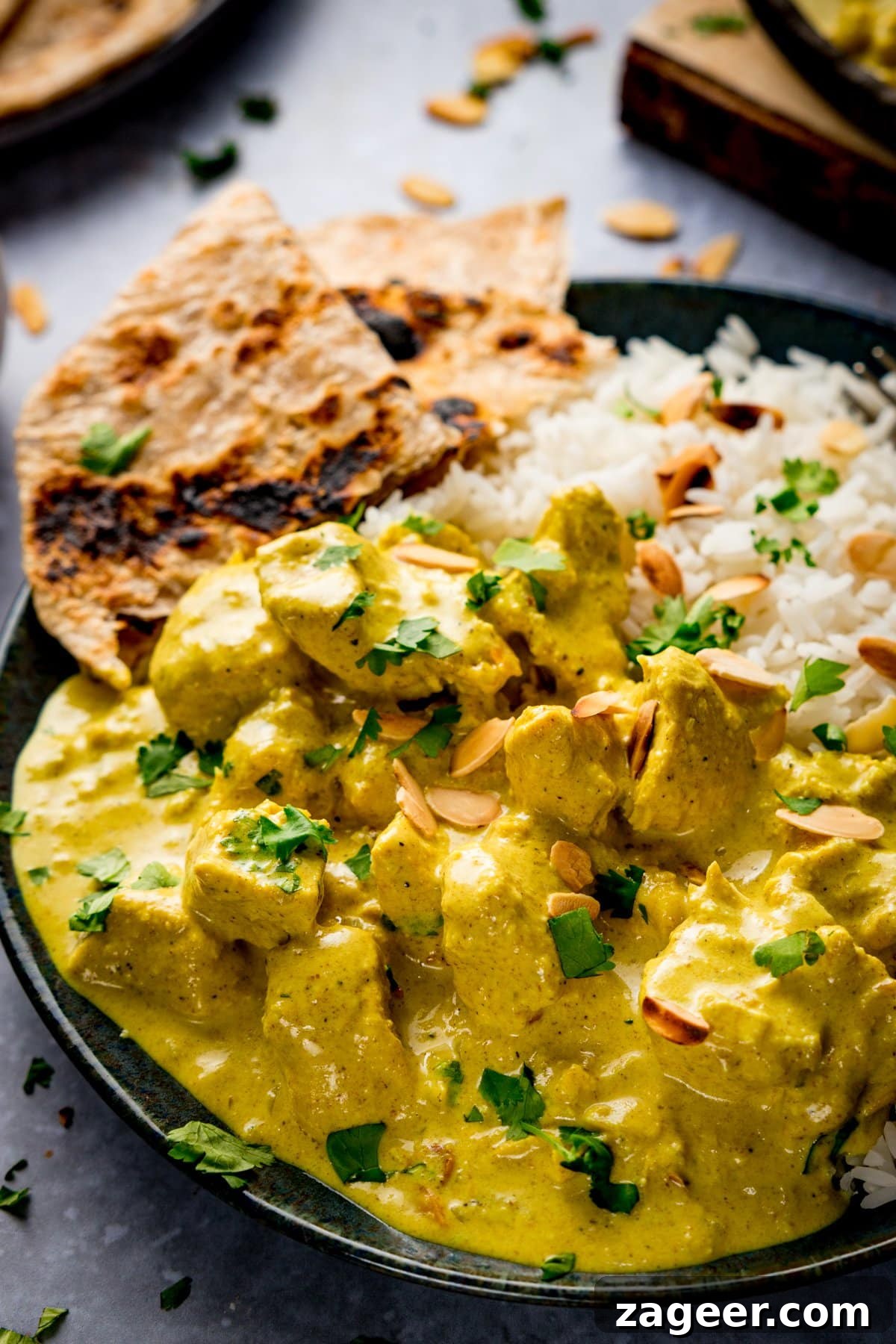 Creamy Chicken Pasanda Recipe: Aromatic Mughlai Curry Guide 13 Dark plate with Chicken pasanda, rice and chapati on. Pasanda has coriander and toasted almonds sprinkled on top.