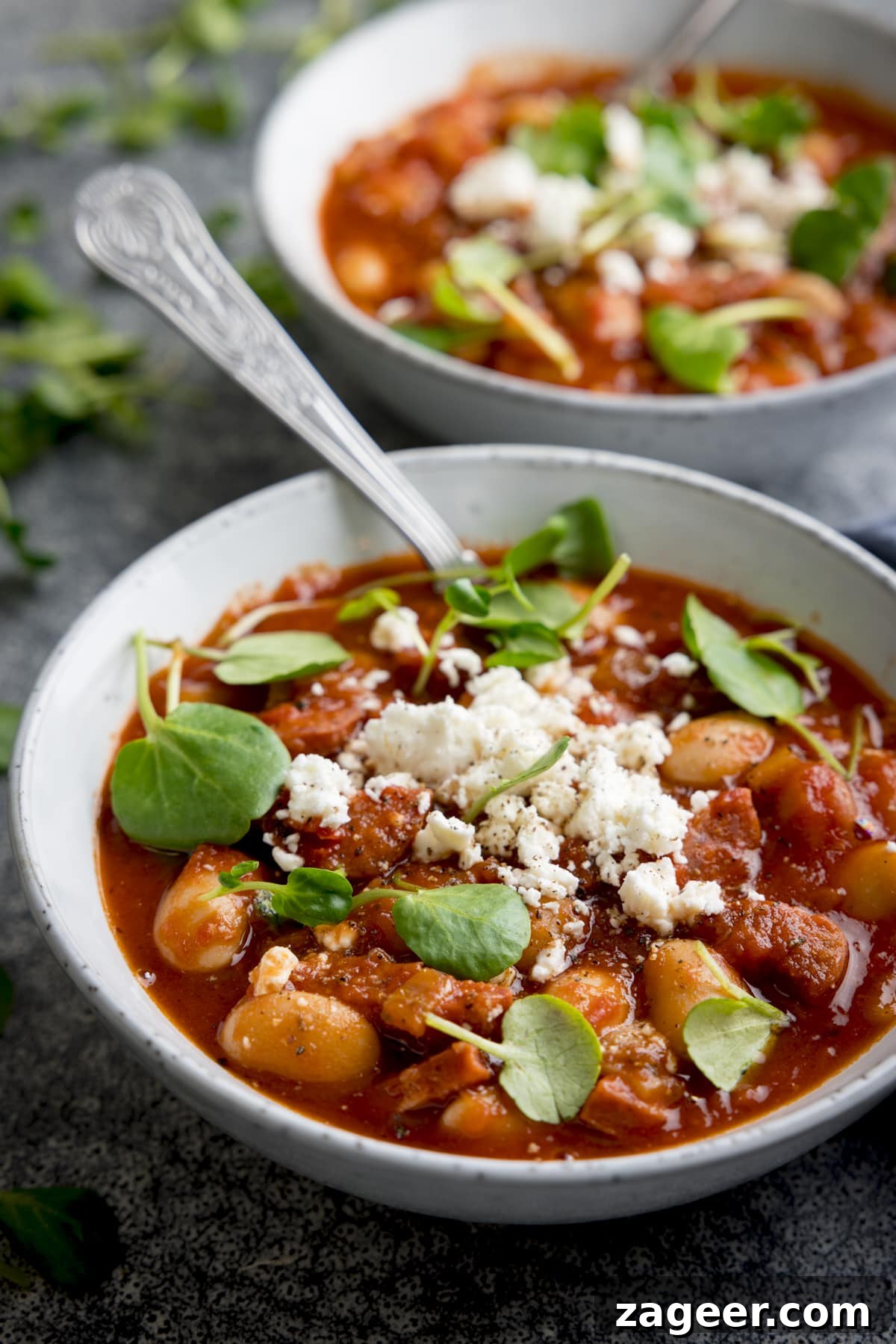 20-Minute Chorizo and Butterbean Soup Recipe 2 Two light grey bowls filled with chorizo and butterbean soup, topped with feta and watercress. There is a spoon sticking out of the front bowl of soup.