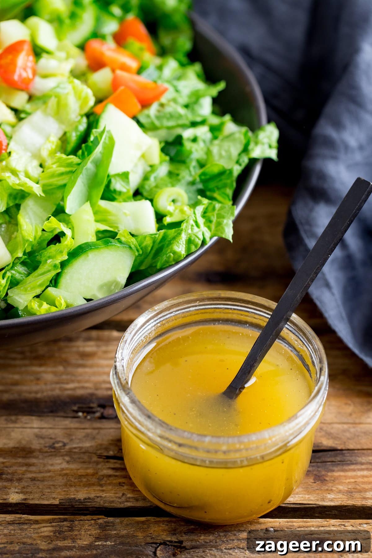 Homemade Honey Mustard Dressing Recipe 2 Jar of honey mustard salad dressing with a black spoon sticking out on a wooden table. Bowl of salad in the background.