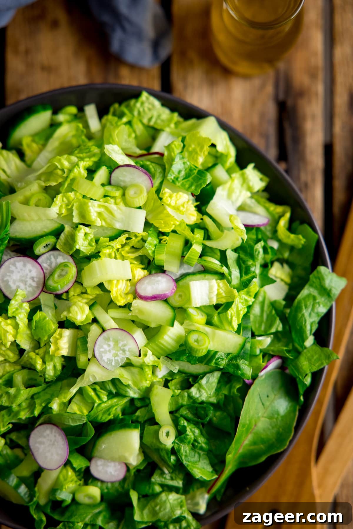 Crisp Green Salad with Tangy Vinaigrette Dressing 4 Overhead image of a bowl of simple green salad on a wooden table. Salad servers, salad dressing and a napkin are also in shot.