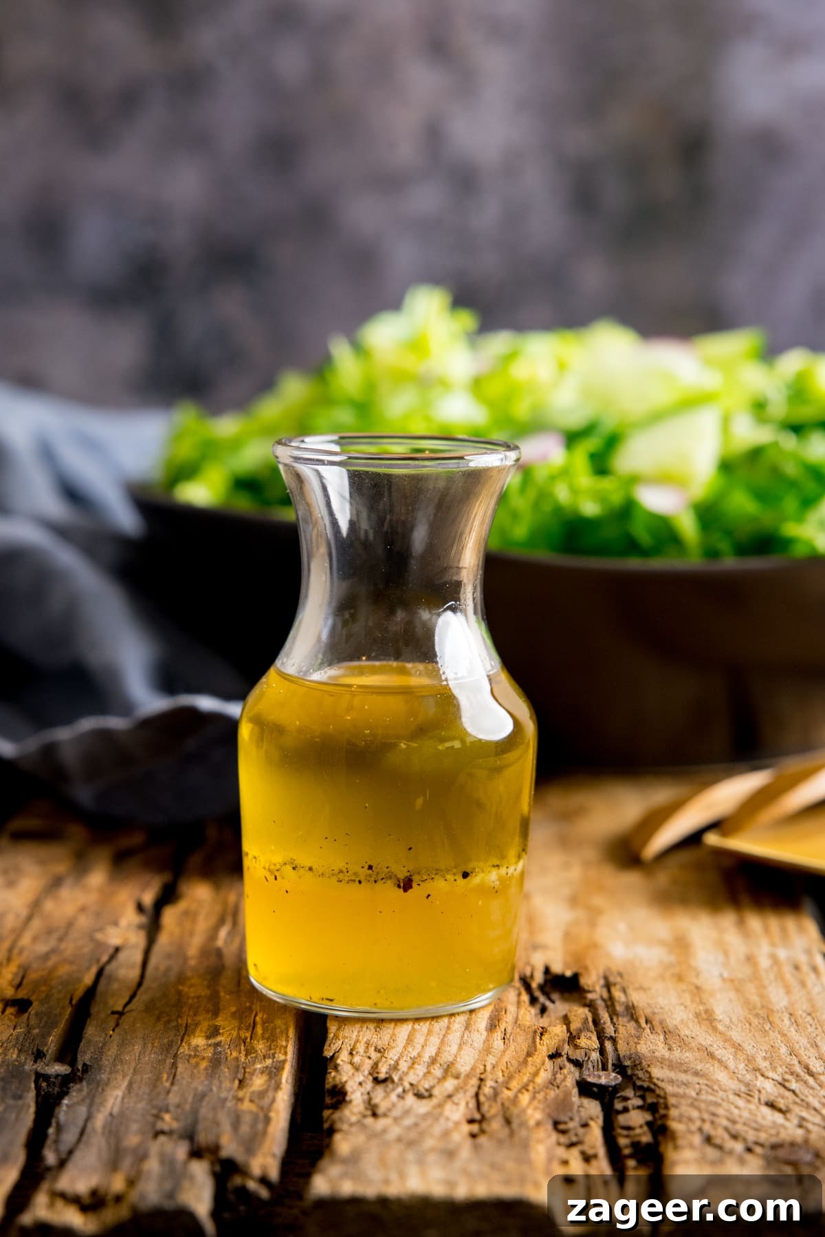 Crisp Green Salad with Tangy Vinaigrette Dressing 5 Small glass jar filled with vinaigrette dressing on a wooden table. In the background is a bowl of salad and a blue napkin.