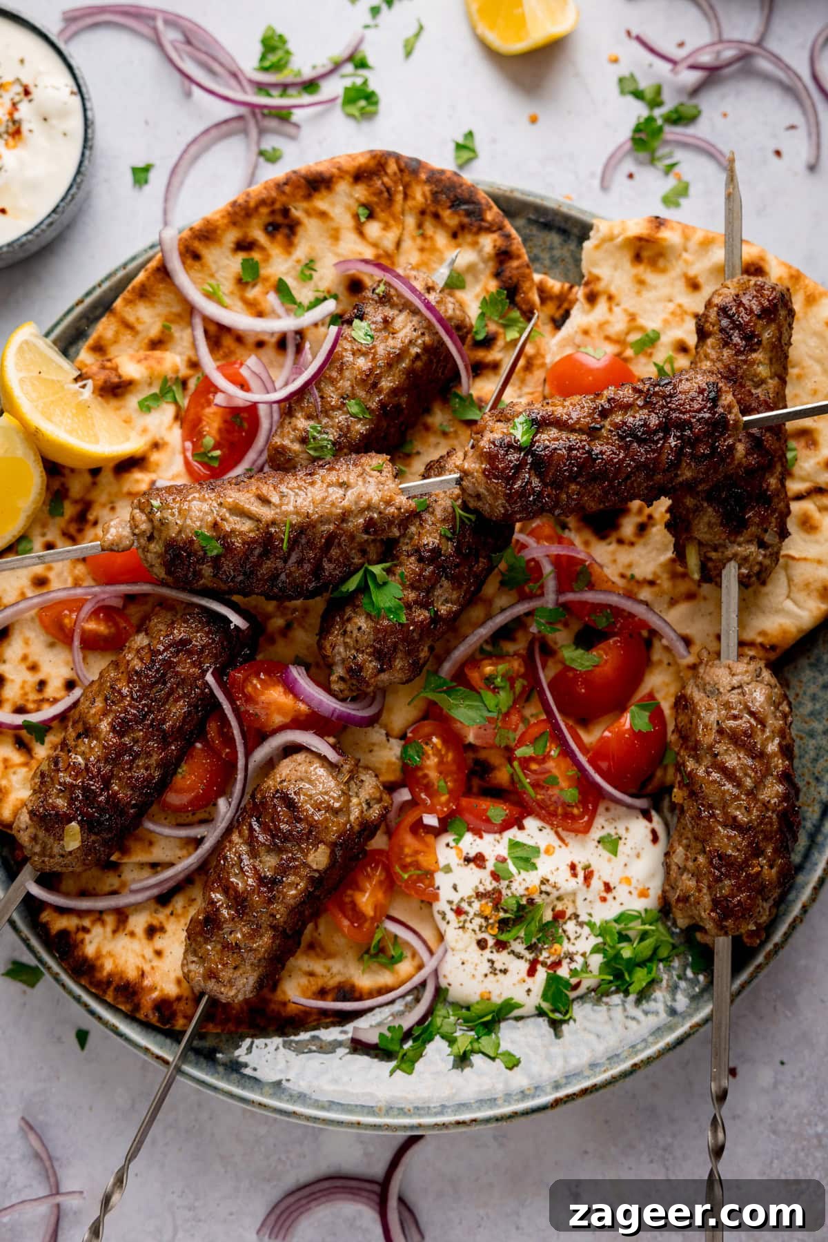 Authentic Lamb Kofta Recipe with Tangy Yogurt Sauce 2 Overhead tall image of grilled lamb kofta skewers on flatbreads, on a plate, with sliced tomatoes, red onions, lemon wedges, fresh herbs, and a side of whipped feta.