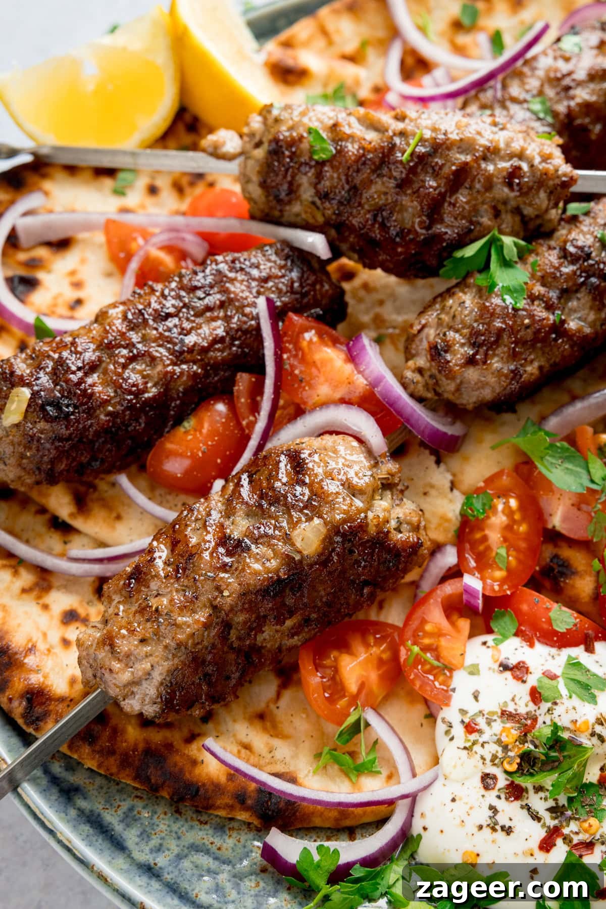 Authentic Lamb Kofta Recipe with Tangy Yogurt Sauce 7 Tall close-up image of grilled lamb kofta skewers on flatbreads, on a plate, with sliced tomatoes, red onions, lemon wedges, fresh herbs, and a side of whipped feta.