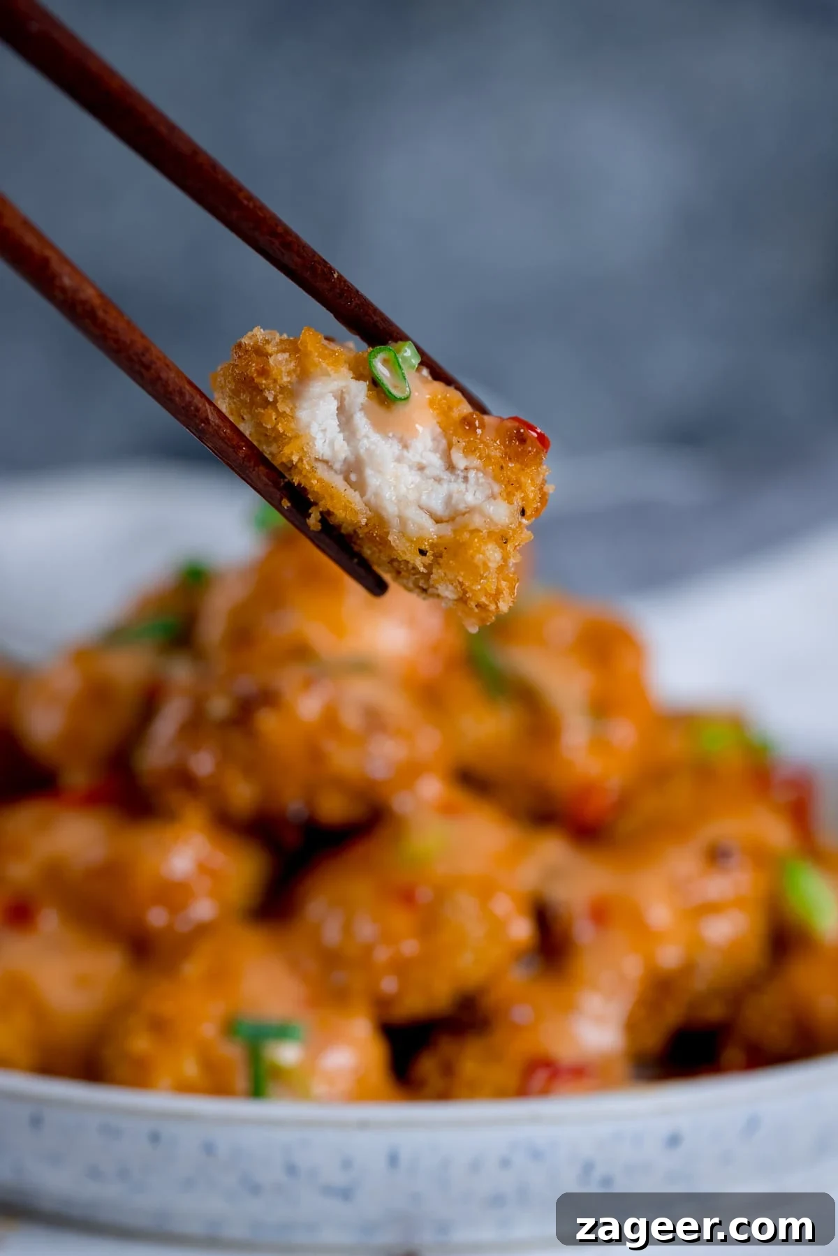 Sichuan Bang Bang Chicken Salad Recipe: Spicy Sesame Chicken 4 Piece of bang bang chicken with a bite out being held with a pair of chopsticks. Bowl of bang bang chicken in the background.