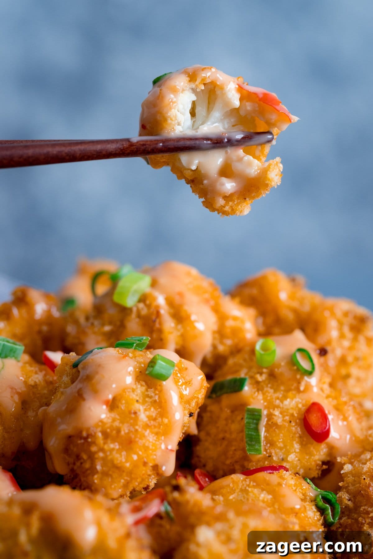 Crispy Bang Bang Cauliflower with Spicy Creamy Sauce 5 Piece of bang bang cauliflower with a bite out of it being lifted by chopsticks from a plate of bang bang cauliflower
