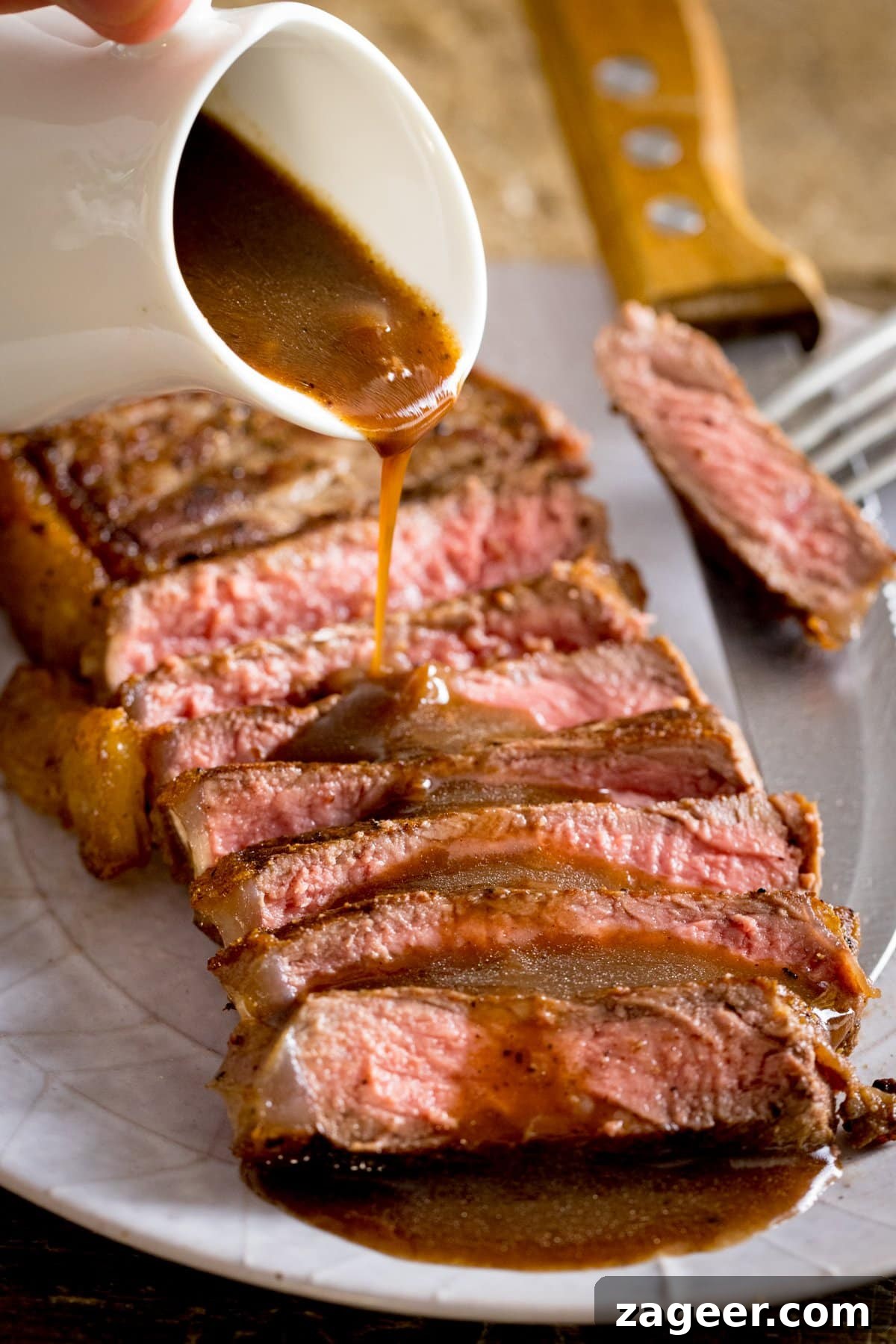 Rich Red Wine Jus Recipe for Steaks and Roasts 2 A plate of cooked sliced steak with red wine just being poured over the top from a white jug, a fork with a piece of cooked steak on the side of the plate