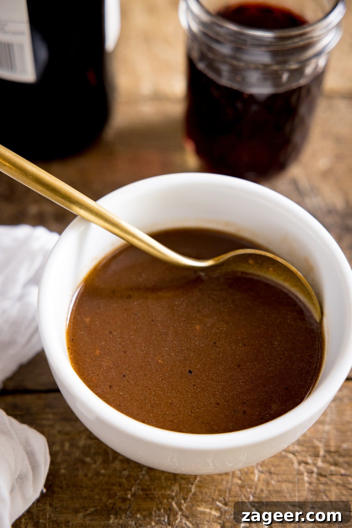 Rich Red Wine Jus Recipe for Steaks and Roasts 4 A tall image of a white bowl of red wine jus being stirred with a spoon on a wooden table, with jars in the background and a tea towel