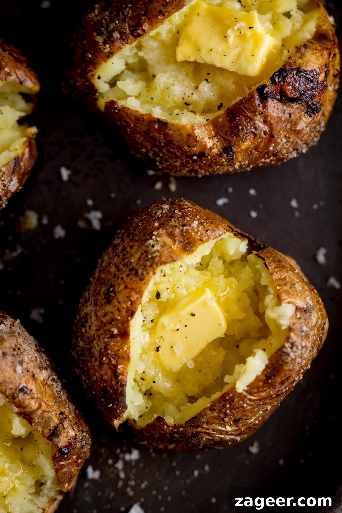 Perfectly Crispy Baked Potato: Foolproof Oven Method 2 Overhead of an open, butter-filled baked potato on a dark surface. Further baked potatoes surround the main one.