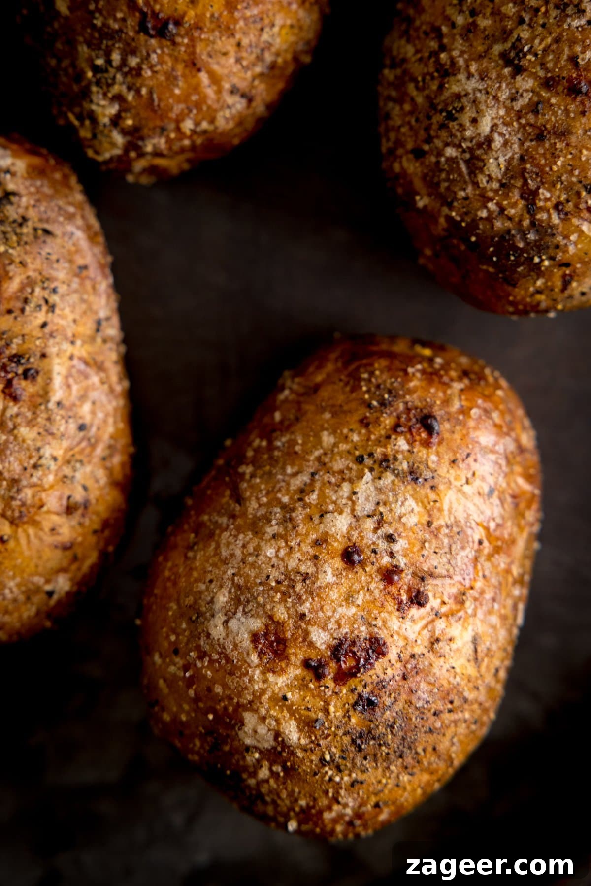 Perfectly Crispy Baked Potato: Foolproof Oven Method 4 Overhead of an unopened baked potato on a dark tray. Further baked potatoes are arranged around the main one.