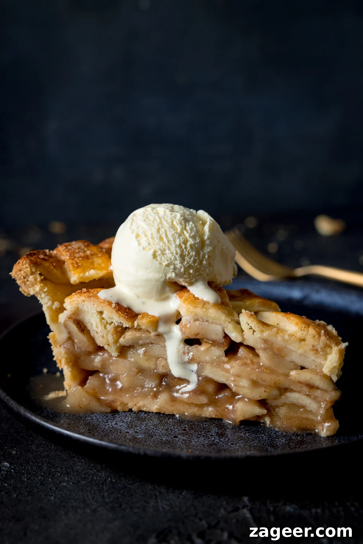 Ultimate Deep-Dish Apple Pie Recipe for Perfect Crust and Filling 2 A side shot of a slice of deep dish apple pie with a scoop of vanilla ice cream on top, on a blue plate with a napkin and fork in the background on a grey table.