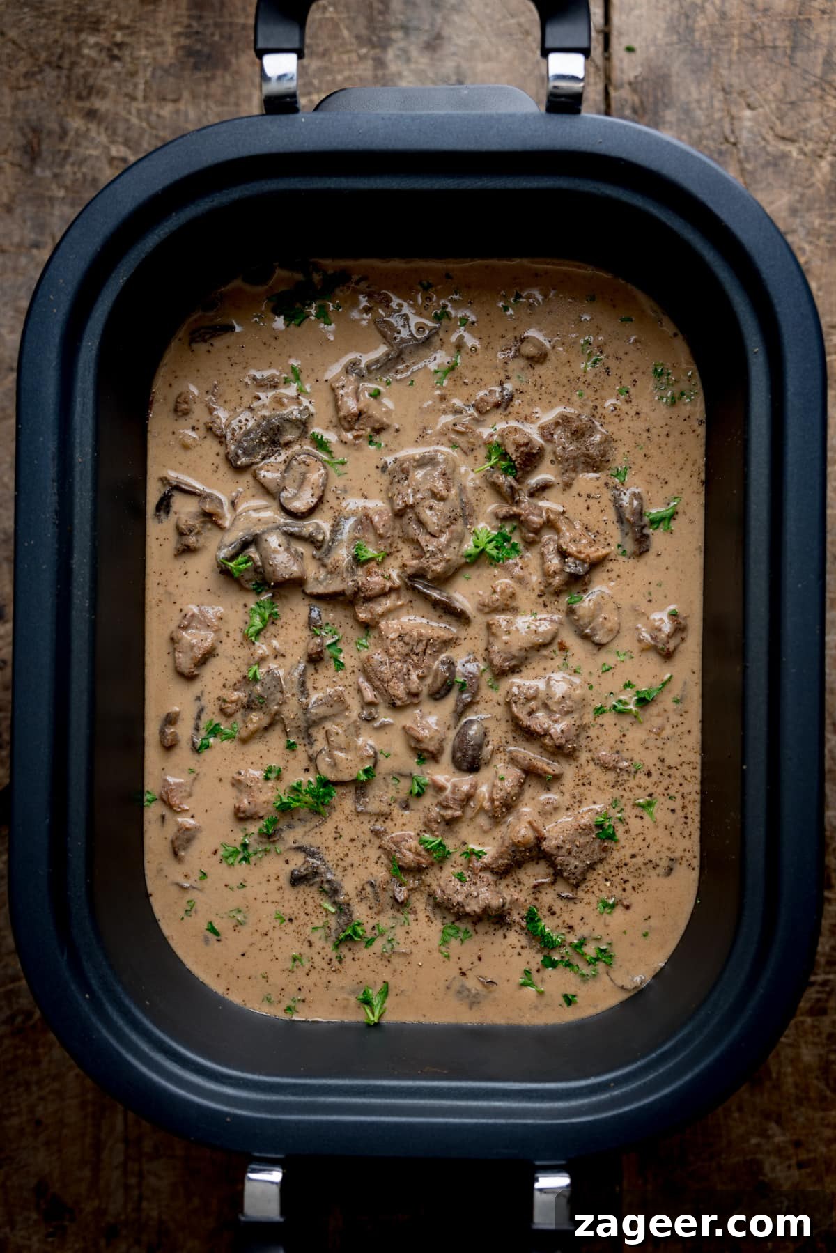 Overhead image of slow cooked beef stroganoff in a slow cooker, on a wooden table. There is parsley sprinkled on top.