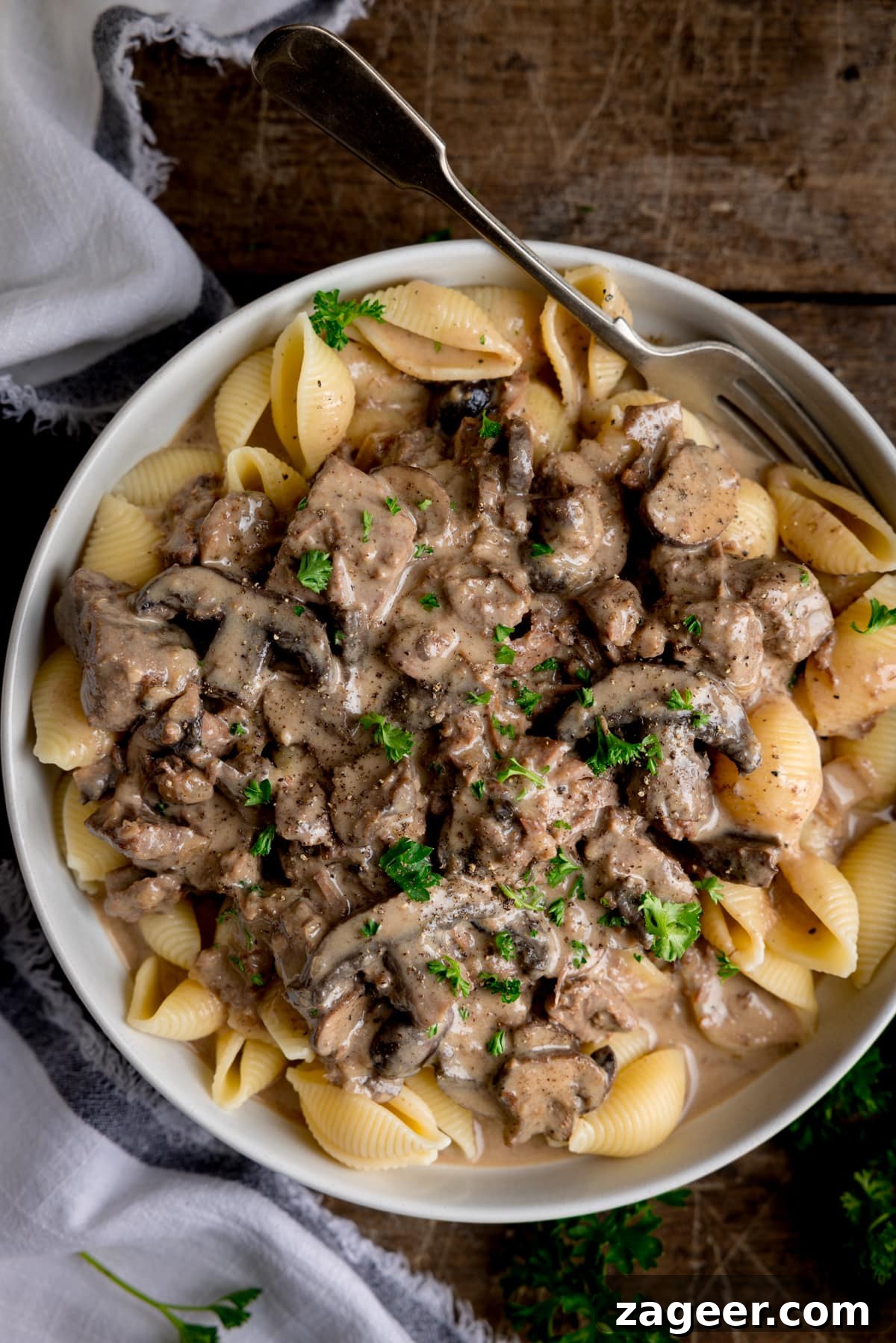 White bowl filled with slow cooker beef stroganoff served over pasta shells. Parsley is sprinkled on top.