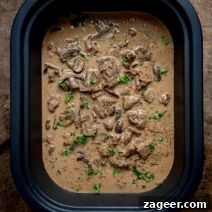 Overhead square image of slow cooked beef stroganoff, with parsley on top, in a slow cooker, on a wooden table.