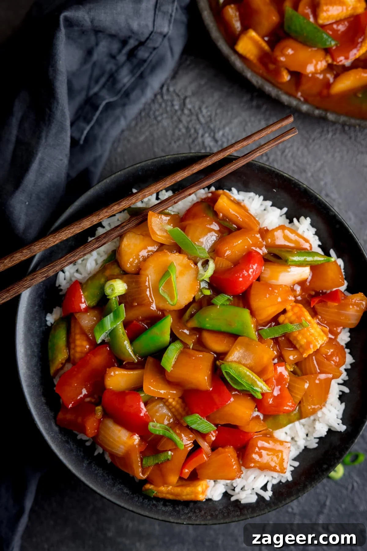 Sweet and Sour Vegetable Stir-Fry Recipe for Tangy Flavor 2 Sweet and sour vegetables on a bed of rice in a black bowl on a dark surface. There is a further bowl of sweet and sour vegetables just in shot at the top of the image.