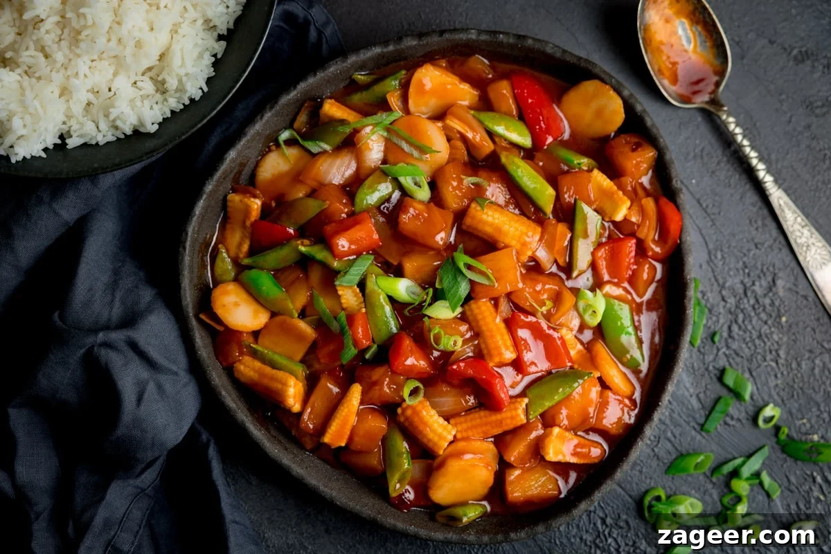 Sweet and Sour Vegetable Stir-Fry Recipe for Tangy Flavor 4 Overhead image of sweet and sour vegetables in a black serving bowl. There is a spoon, navy napkin, chopped springs onions and bowl of rice around the bowl of sweet and sour vegetables.