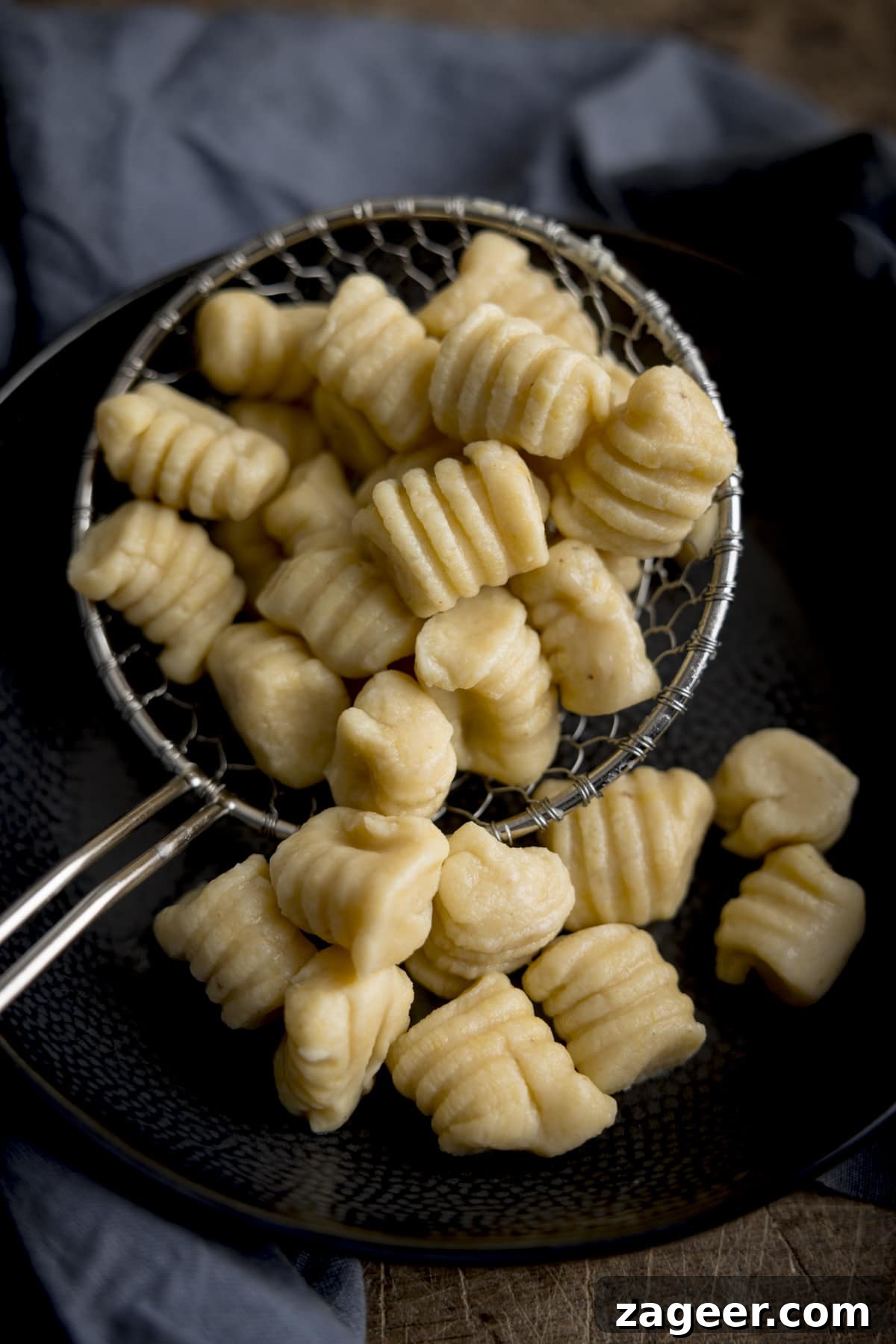 Authentic Homemade Potato Gnocchi Recipe for Fluffy Results 6 Homemade gnocchi in a slotted spoon, being added to a dark bowl. There is a blue napkin surrounding the bowl.