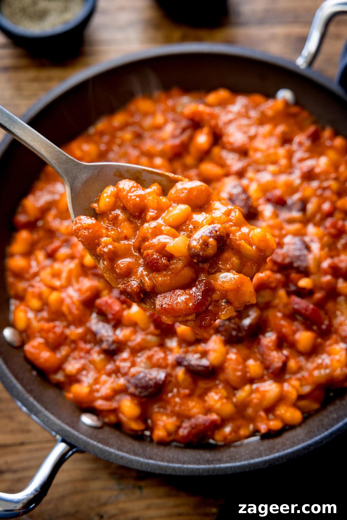 Smoky Barbecue Baked Beans Recipe for Summer Cookouts 2 A tall shot of BBQ Beans. The BBQ Beans are in a black skillet, with a silver spoon sticking out of the dish. In the top of the background, you can see two small black dishes, one with salt and one with ground pepper. This is all set on a wooden background.