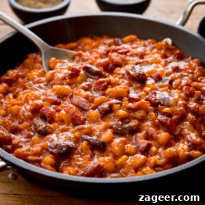 Smoky Barbecue Baked Beans Recipe for Summer Cookouts 17 A square shot of BBQ Beans. The BBQ Beans are in a black skillet, with a silver spoon sticking out of the dish. In the top of the background, you can see two small black dishes, one with salt and one with ground pepper. This is all set on a wooden background.