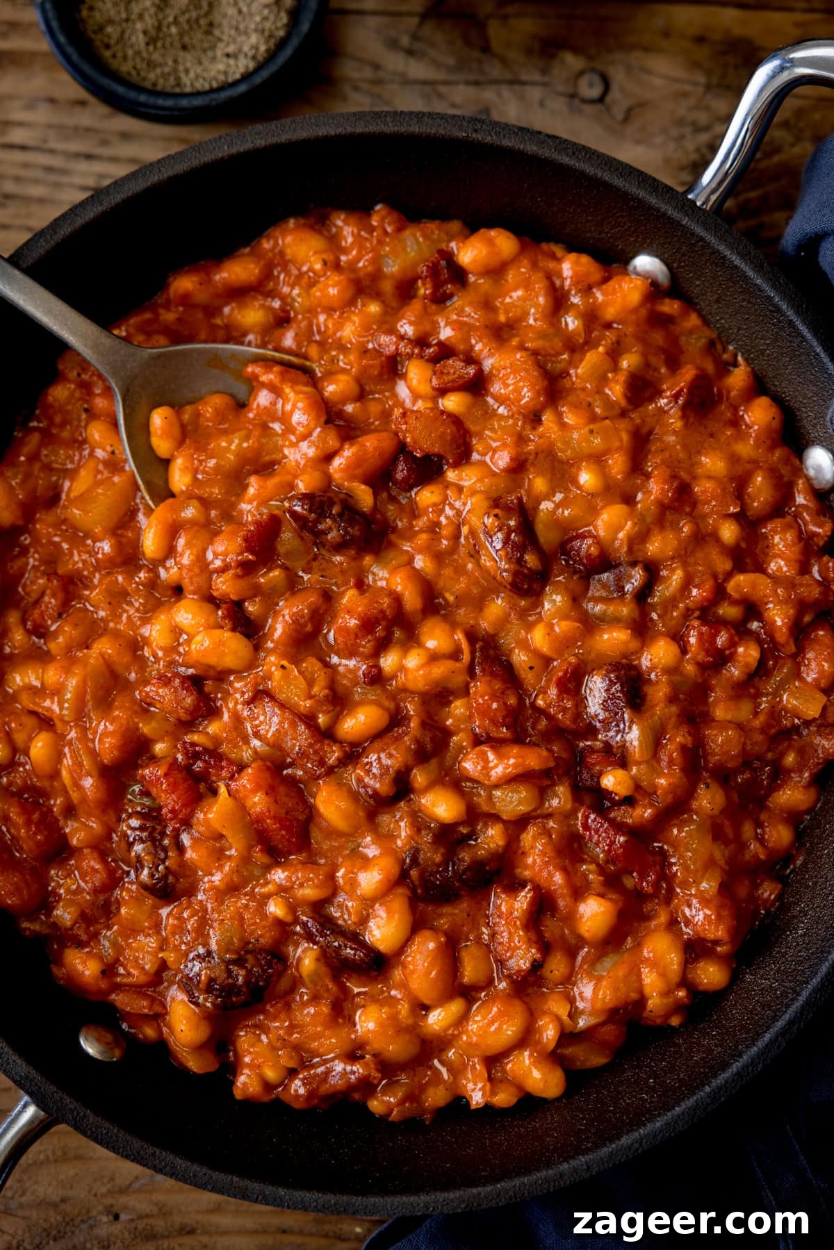 Smoky Barbecue Baked Beans Recipe for Summer Cookouts 4 A tall, overhead shot of BBQ Beans. The BBQ Beans are in a black skillet, with a silver spoon sticking out of the dish. In the top of the background, you can see two small black dishes, one with salt and one with ground pepper. This is all set on a wooden background.