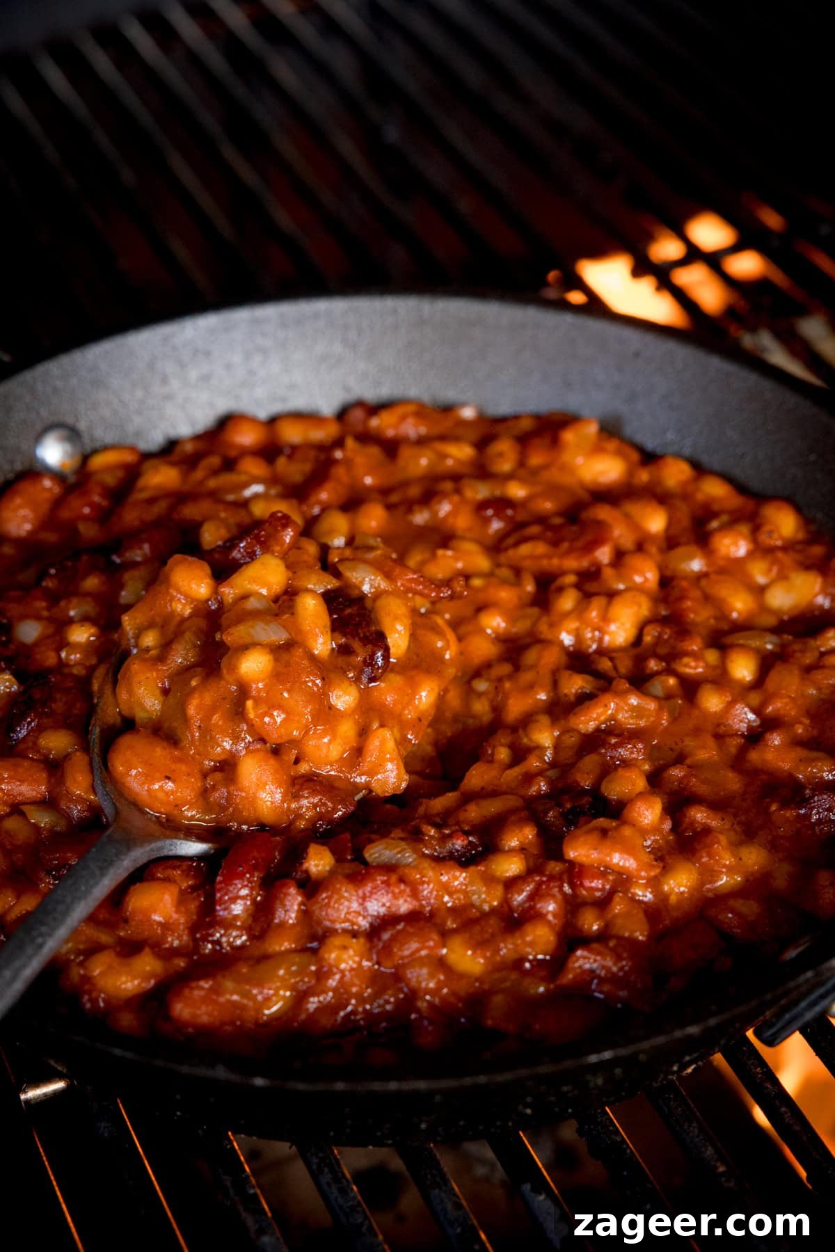 Smoky Barbecue Baked Beans Recipe for Summer Cookouts 5 A square shot of BBQ Beans. The BBQ Beans are in a black skillet, with a silver spoon sticking out of the dish, which is scooping some of the beans in the dish. In the background, you can see that the skillet is on a lit barbeque, in the top right of the background you can see a small flame from the barbeque.