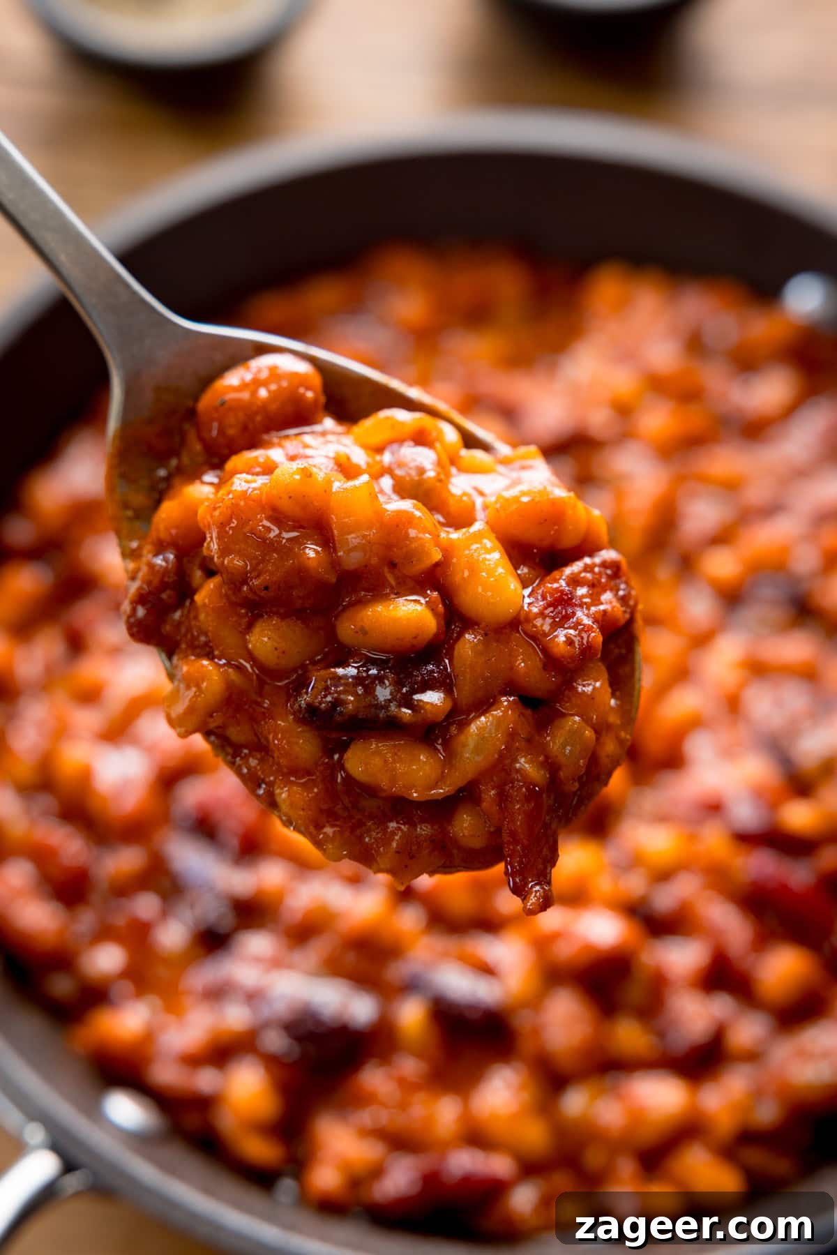 Smoky Barbecue Baked Beans Recipe for Summer Cookouts 10 A tall, overhead shot of BBQ Beans. The BBQ Beans are in a black skillet. In the foreground, there is a silver spoon with some BBQ Beans in it, being held above the skillet of beans. In the top of the background, you can see two small black dishes, one with salt and one with ground pepper. This is all set on a wooden background.