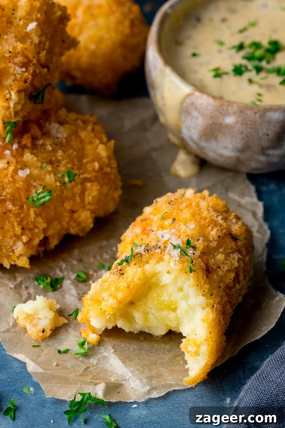 Cheesy Baked Potato Croquettes: Crispy Oven-Baked Recipe 6 potato croquette with a bit taken out on a piece of parchment paper. Further potato croquettes and dip in the background.