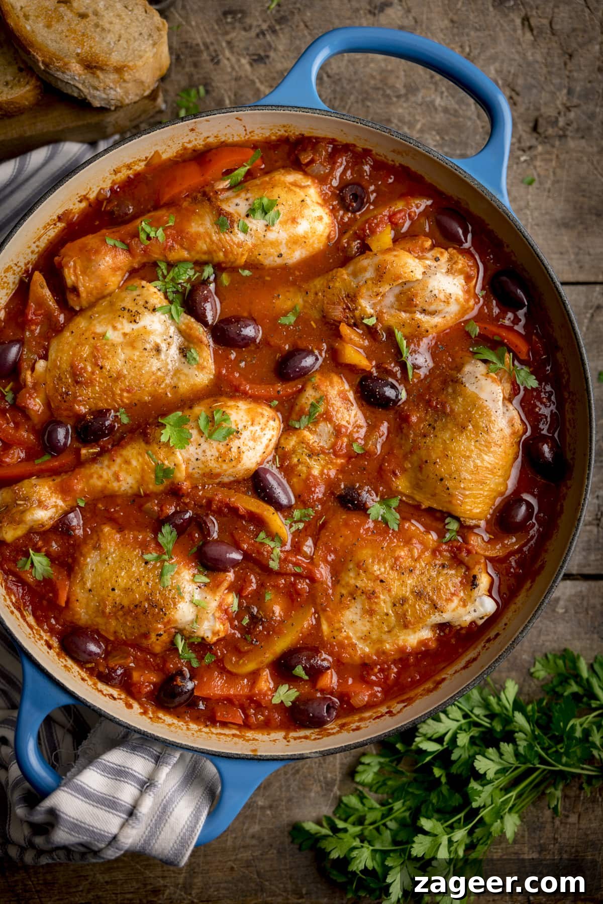 Classic Chicken Cacciatore Recipe: Rustic Italian Hunter’s Chicken 2 Overhead image of chicken cacciatore in a large, shallow blue casserole dish. The dish is on a wooden table, next to some fresh herbs and sliced bread.