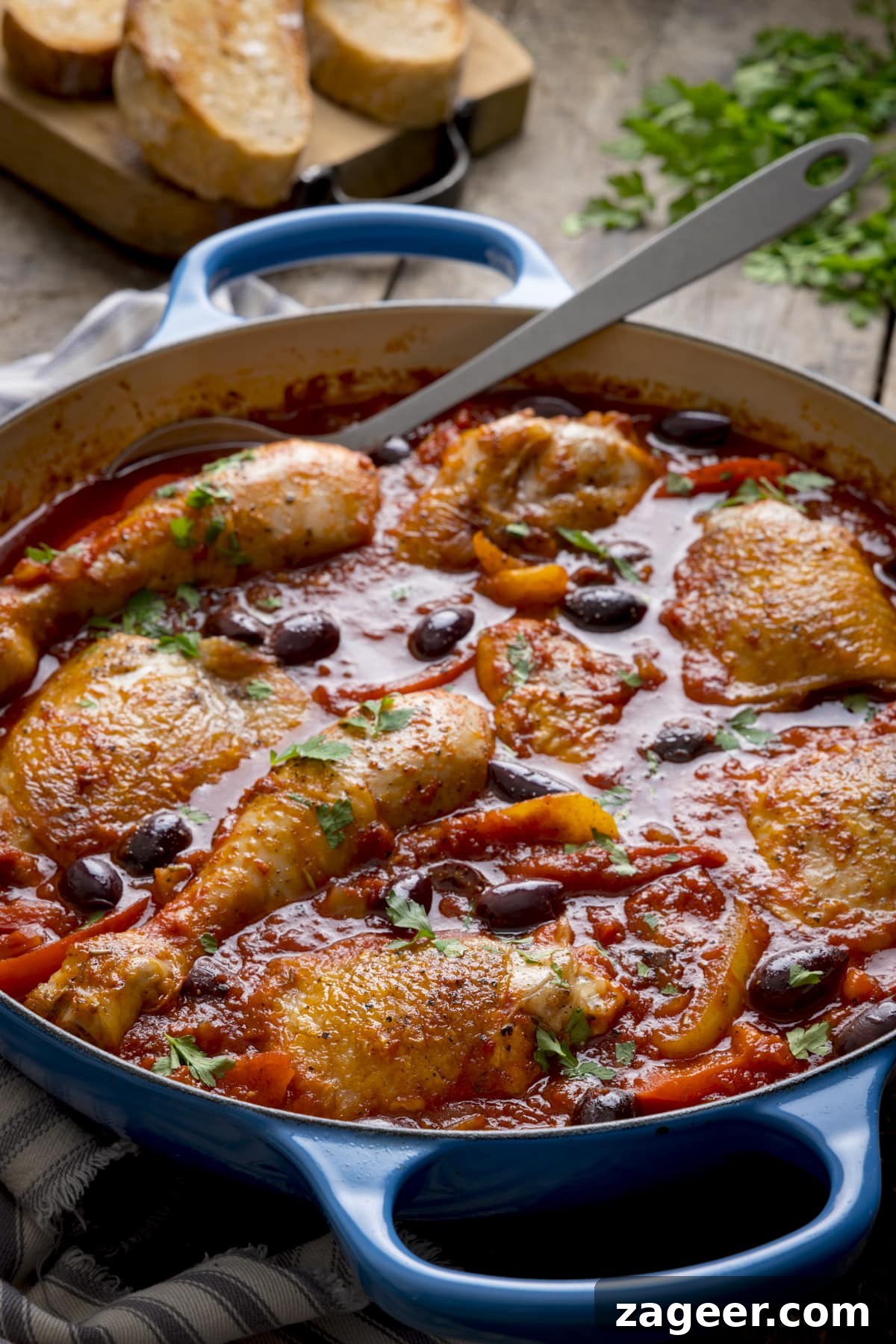 Classic Chicken Cacciatore Recipe: Rustic Italian Hunter’s Chicken 4 Tall image of chicken cacciatore in a large, shallow blue casserole dish, with a silver serving spoon sticking out. The dish is on a wooden table, next to some fresh herbs and sliced bread.