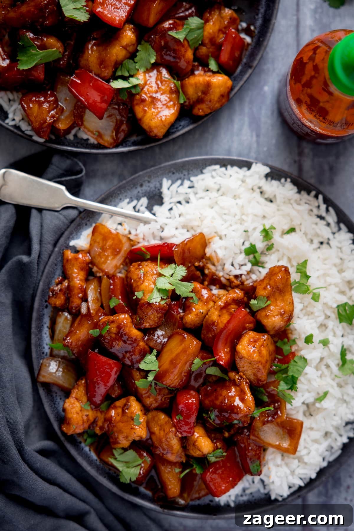 Sticky Pineapple Chicken Recipe with Sweet Tangy Glaze 5 Overhead image of sticky pineapple chicken stir fry and boiled rice on a dark plate with a fork sticking out. The plate is on a blue-grey background, next to a blue-grey napkin. There is a further plate of stir fry and a bottle of sriracha sauce also in shot.