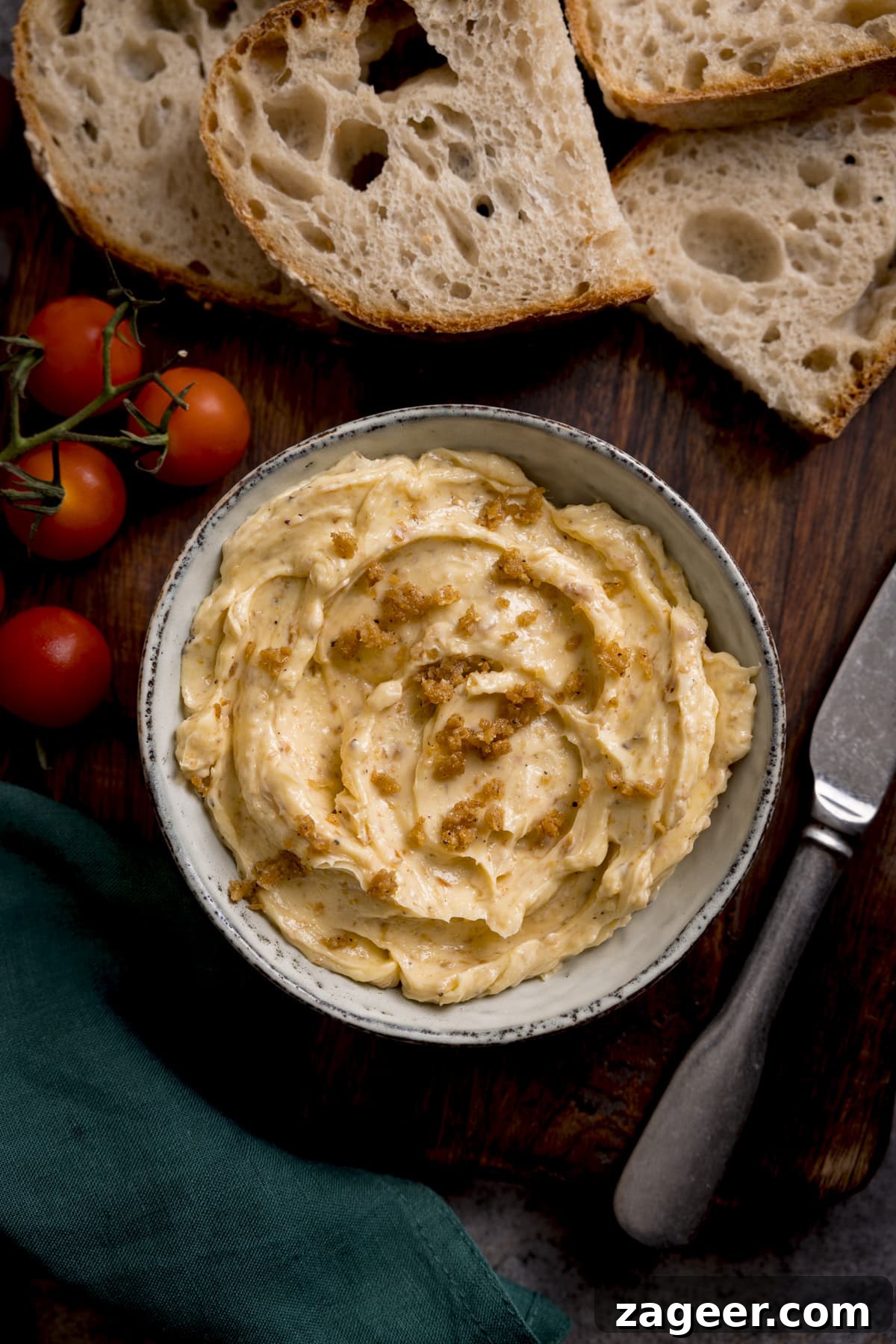 Crispy Chicken Skin Cracklings with Savory Butter Dip 6 Overhead image of chicken skin butter in a grey bowl on a wooden board. There are slices of bread, some vine cherry tomatoes, a knife and a green napkin around the bowl.