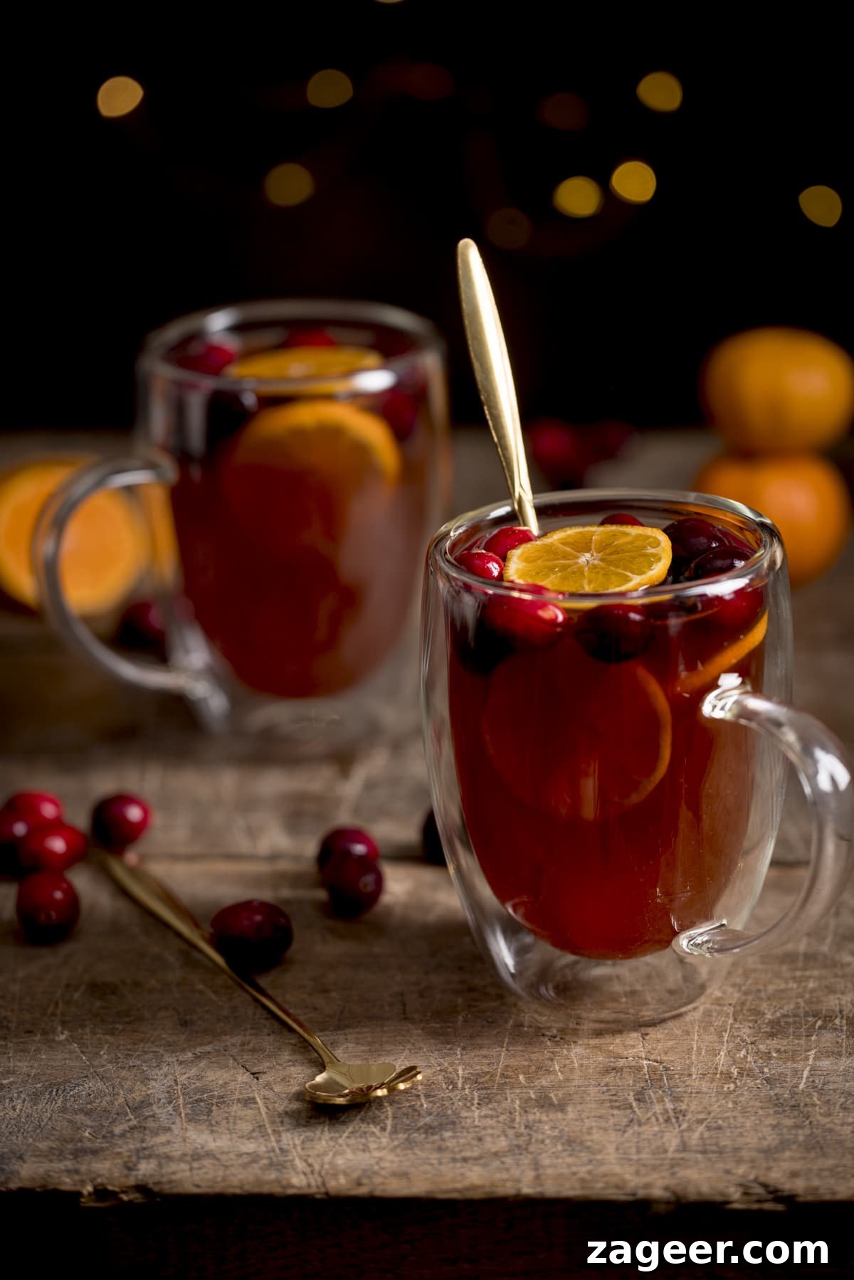 Homemade Mulled Cider Recipe for Cozy Fall Nights 2 2 glass mugs filled with mulled cider. There are clementine slices and cranberries in the cider and the mugs are on a wooden table. The front mug has a gold teaspoon in it.