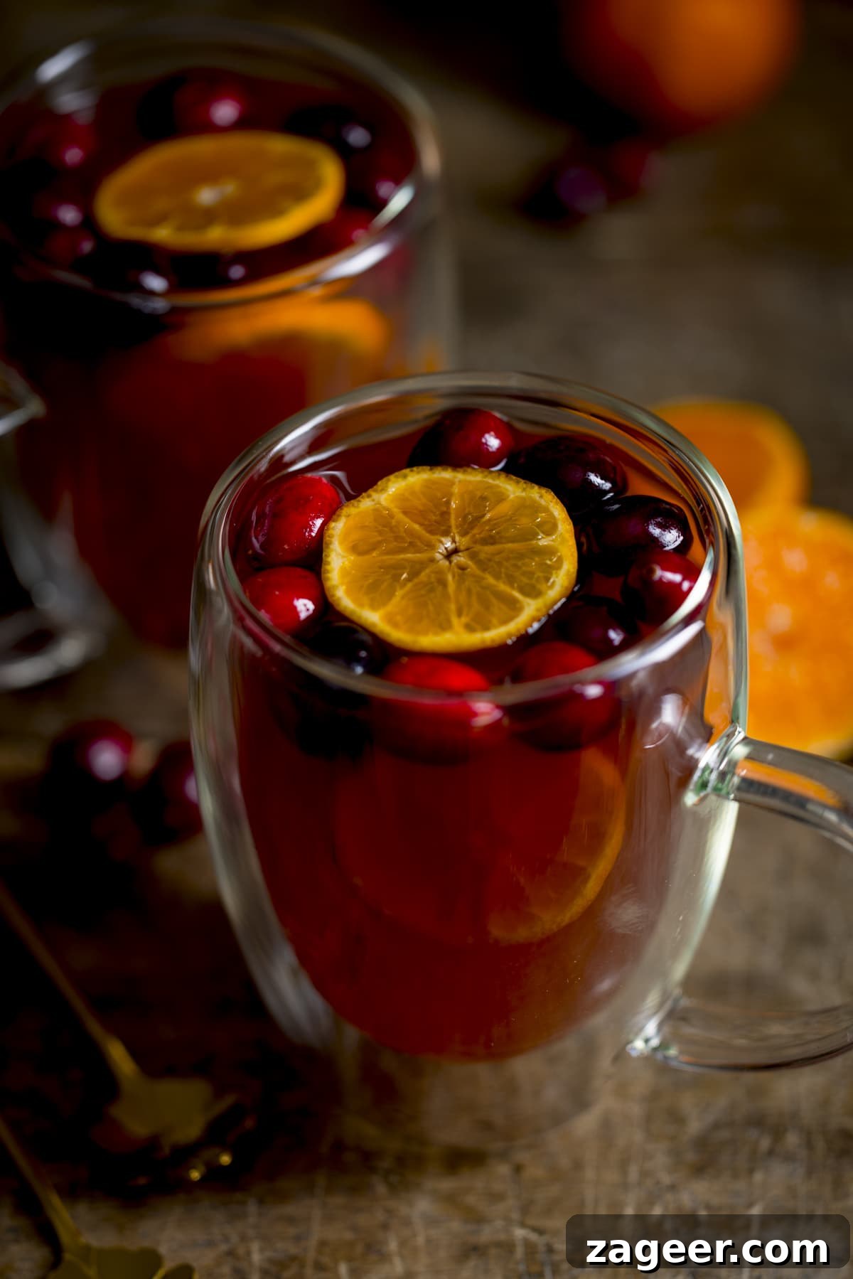 Homemade Mulled Cider Recipe for Cozy Fall Nights 3 2 glass mugs filled with mulled cider. There are clementine slices and cranberries in the cider and the mugs are on a wooden table.