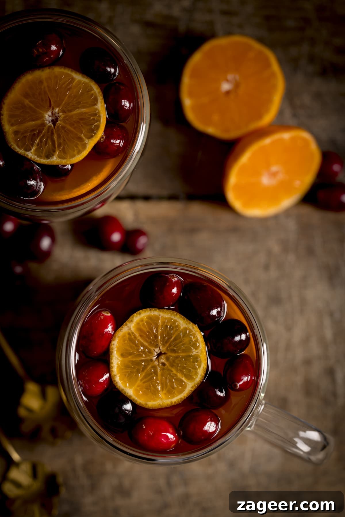 Homemade Mulled Cider Recipe for Cozy Fall Nights 4 Overhead image of two glass mugs filled with mulled cider, cranberries and clementine slices on a wooden table. There is a clementine sliced in half next to the mugs.