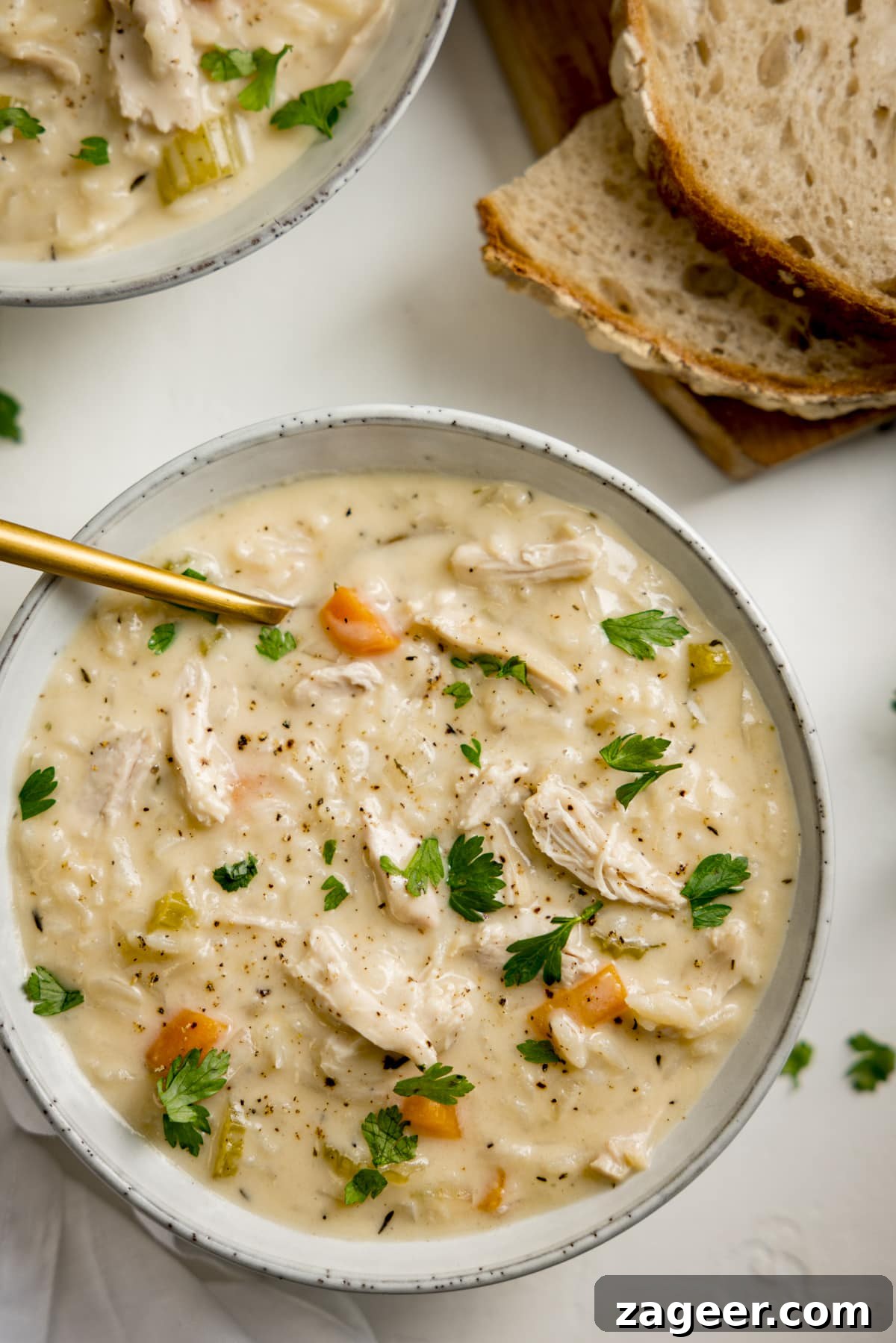 Classic Chicken and Rice Soup Recipe for Comforting Weeknight Meals 8 Overhead image of a light grey bowl filled with chicken and rice soup on a light surface. There is a gold spoon in the soup. There is a further bowl of soup and some slices of bread at the top of the frame.