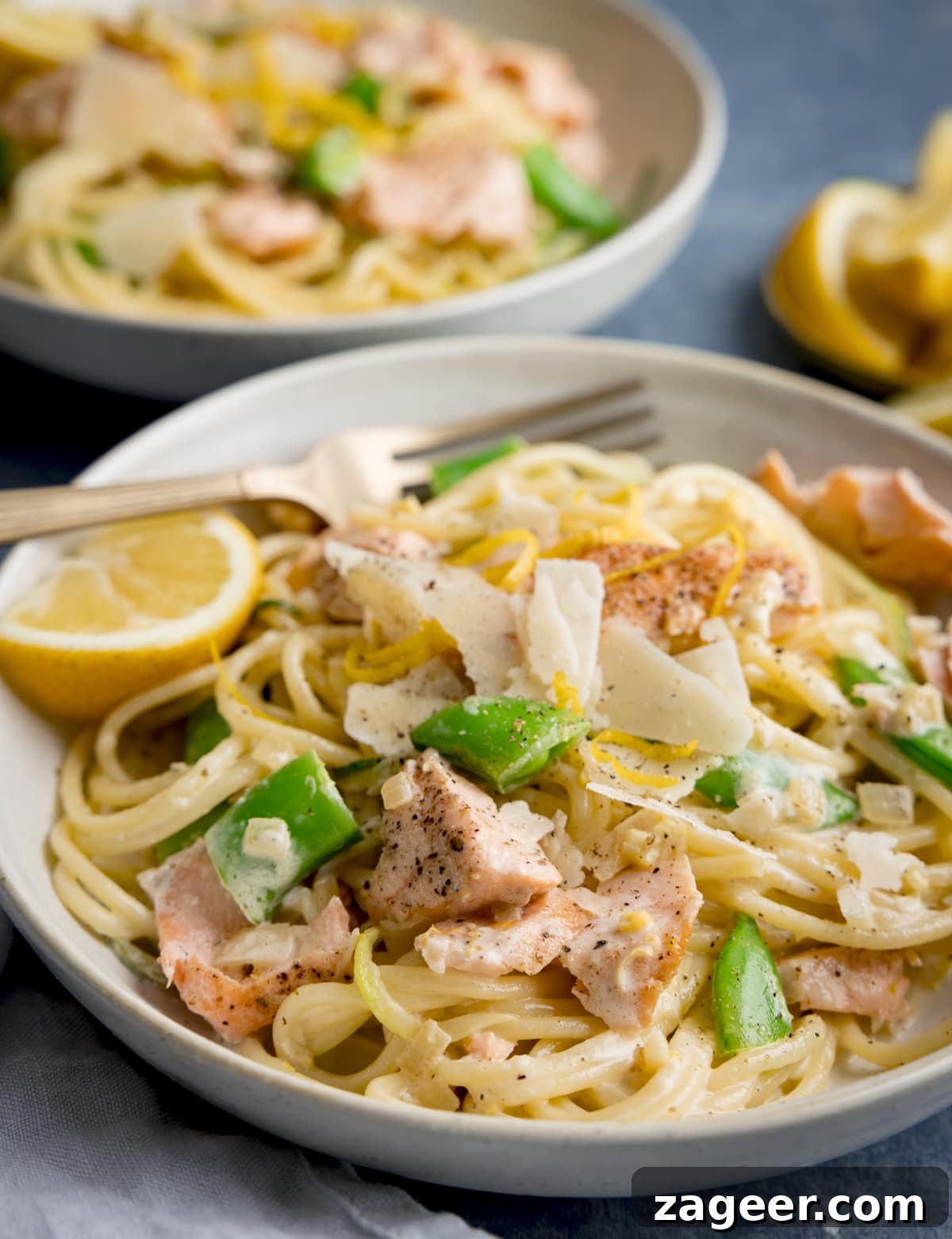 Side on image of salmon and spaghetti in a creamy lemon sauce in a white bowl with a lemon wedge in the bowl. The bowl is on a blue background. There is a further bowl of pasta, a napkin and some lemon wedges nearby.