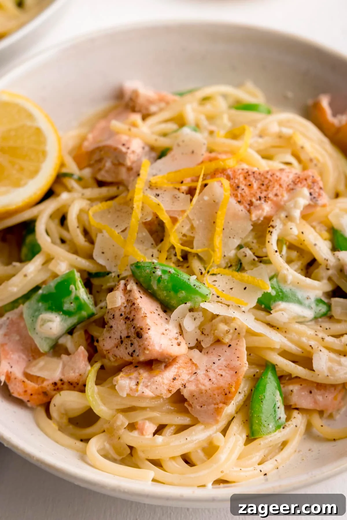 Close up of salmon and spaghetti in a creamy lemon sauce in a white bowl with a lemon wedge in the bowl. The bowl is on a white background.