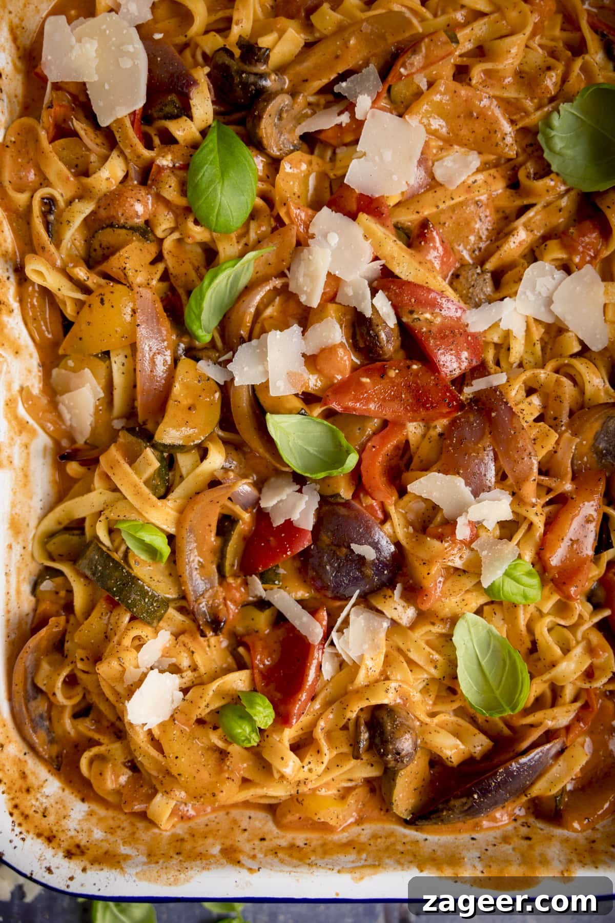 Roasted Veggie Pasta with Garlic and Parmesan 2 Close up of a white and blue roasting tin filled with tagliatelle in a creamy roasted vegetable sauce. There are basil leaves and cheese shavings sprinkled on top.