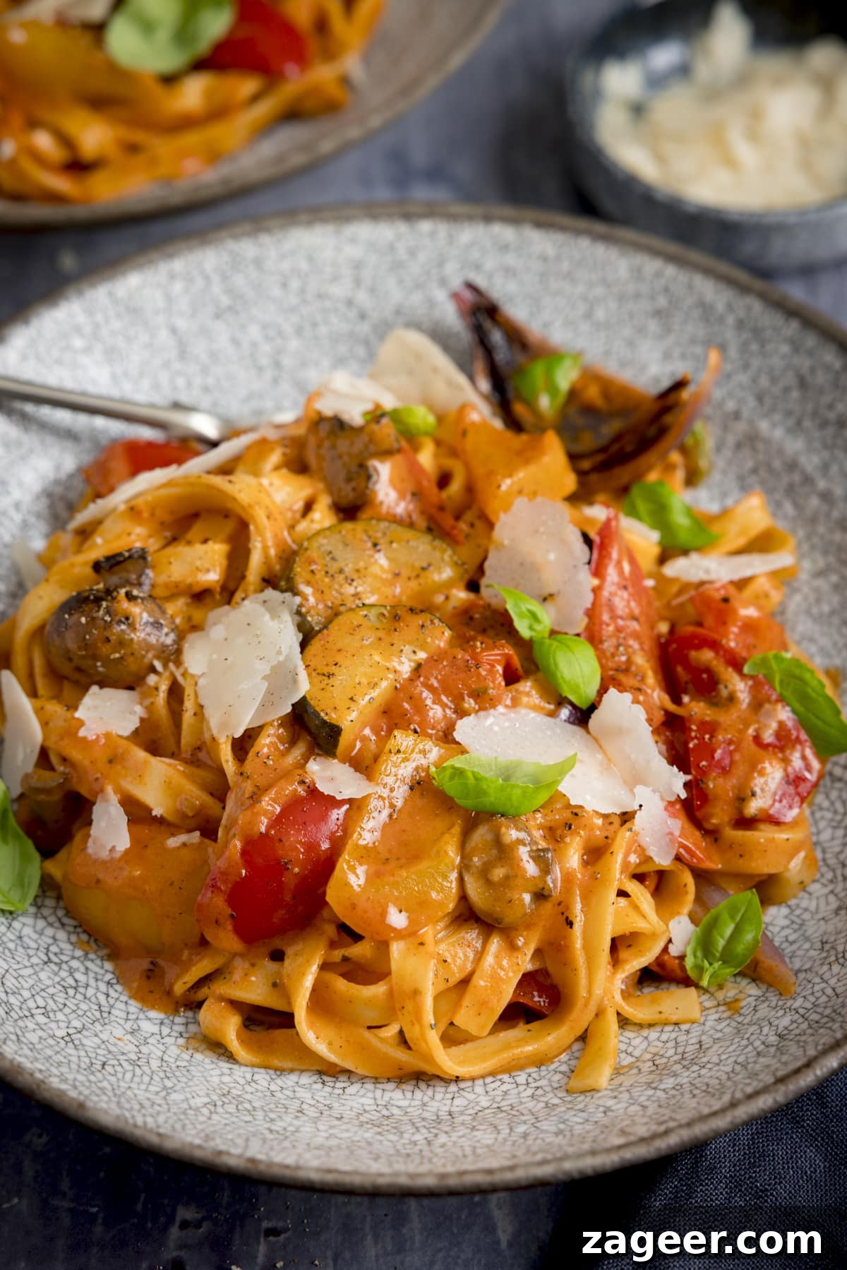 Roasted Veggie Pasta with Garlic and Parmesan 8 Tall image of a bowl of roasted vegetable tagliatelle on a blue background with ingredients scattered around.