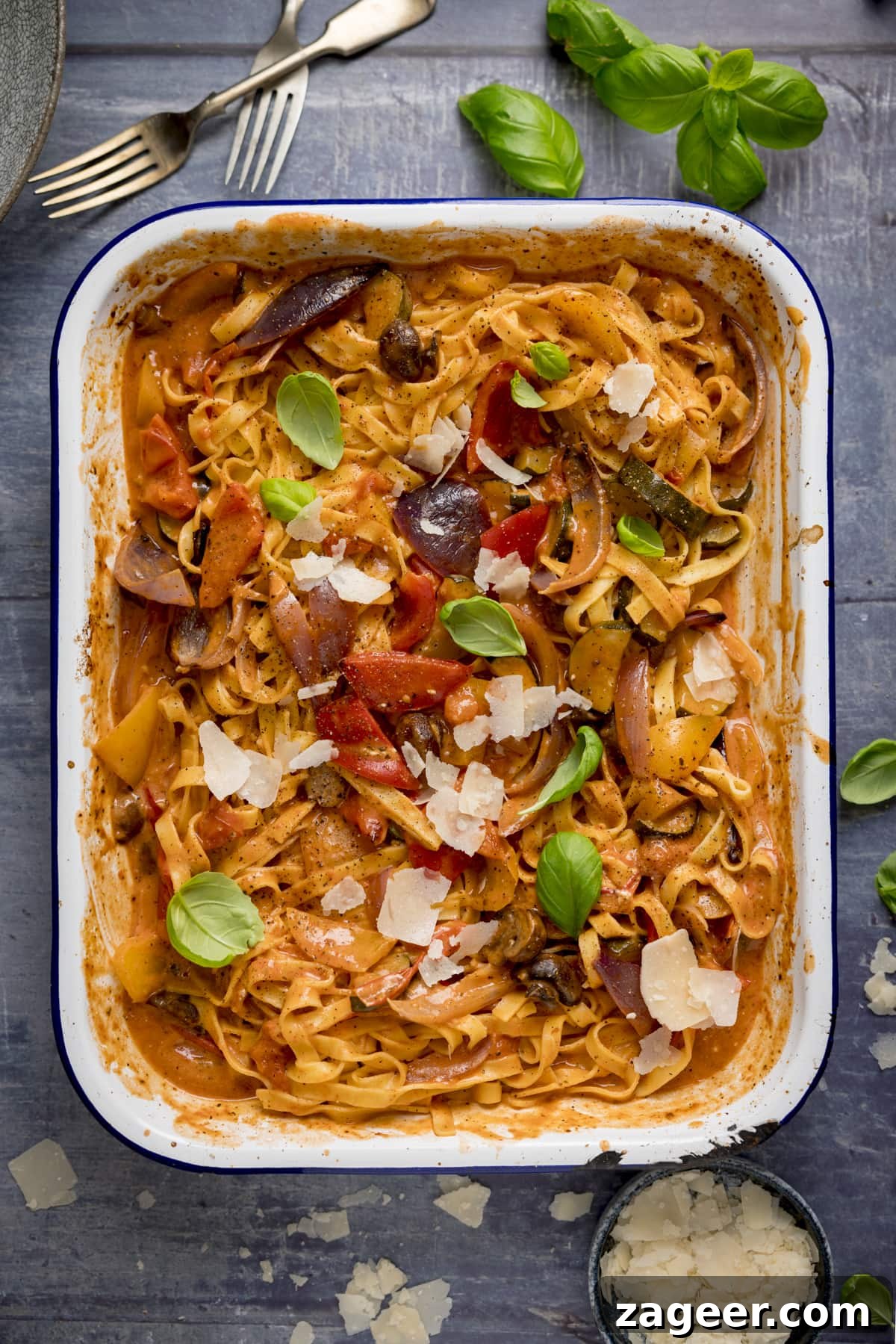 Roasted Veggie Pasta with Garlic and Parmesan 9 A white roasting tin filled with tagliatelle in a creamy roasted vegetable sauce on a blue background. The pasta is topped with a few basil leaves and some cheese shavings.
