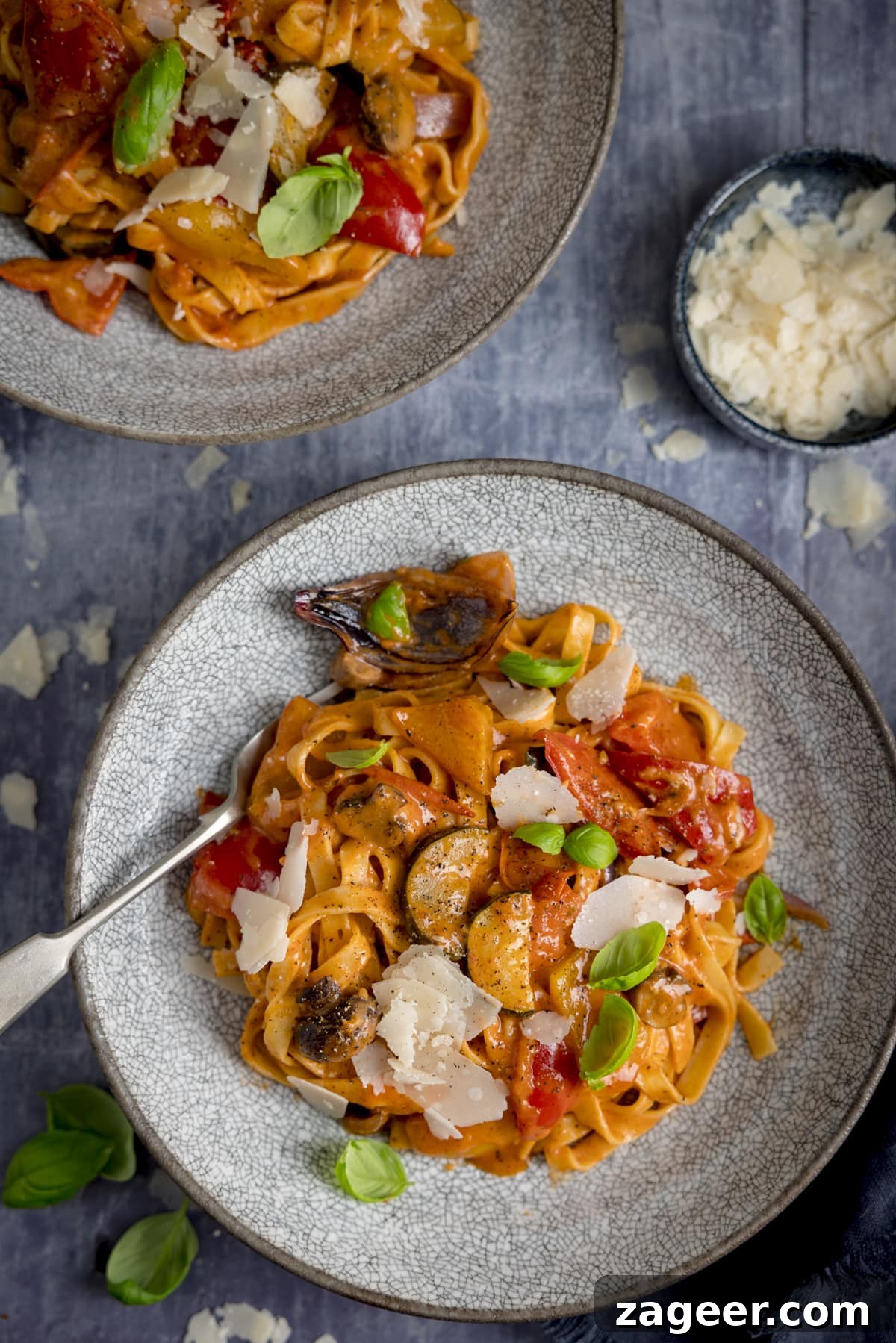 Roasted Veggie Pasta with Garlic and Parmesan 10 Overhead of a bowl of roasted vegetable tagliatelle on a blue background. There is a further bowl of pasta at the top of the frame and a bowl of shaved cheese in shot.