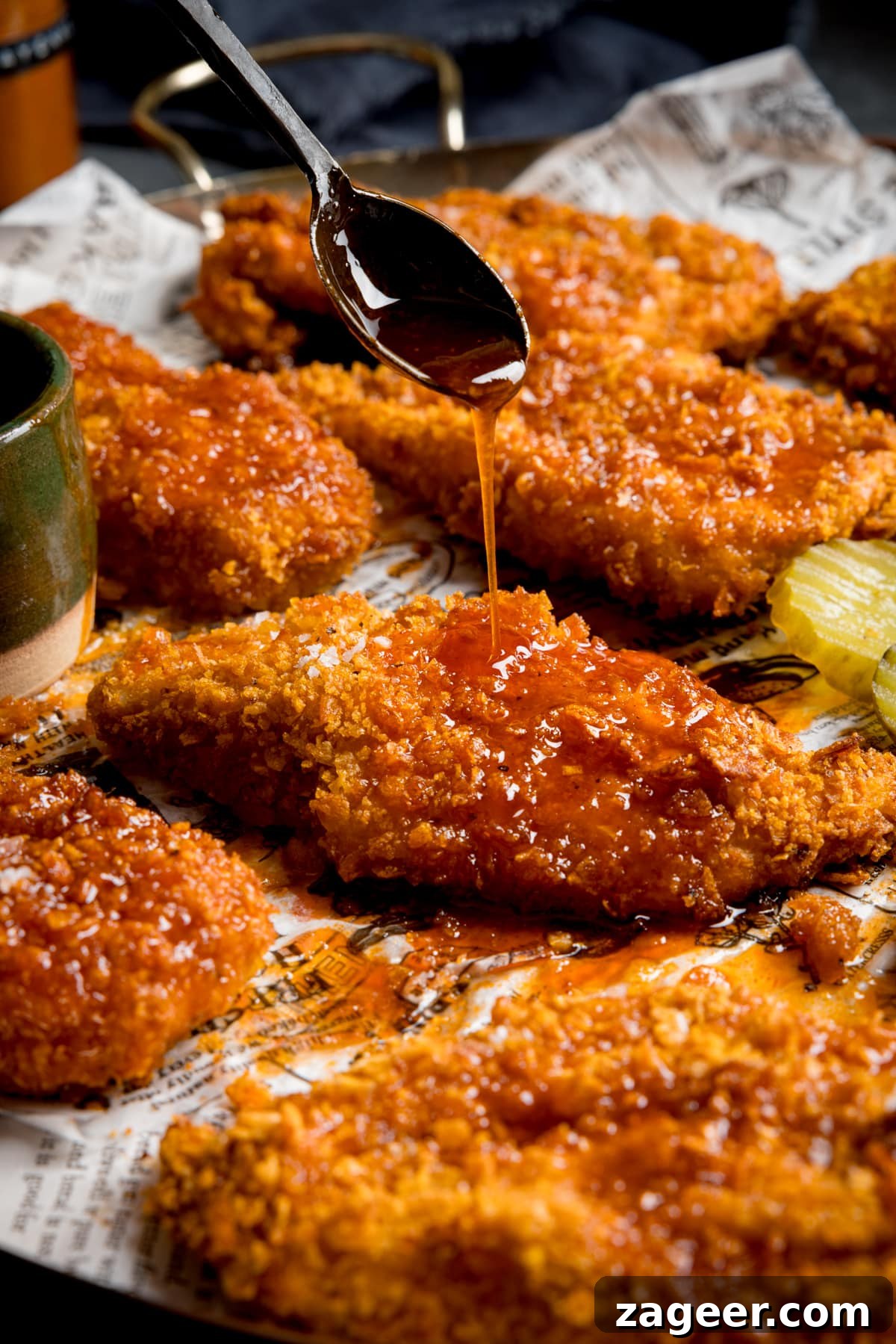 Spicy Honey-Glazed Chicken Recipe 2 Crispy baked chicken breast fillets on a baking tray with sliced pickles. The chicken is being drizzled with hot honey sauce from a black spoon.