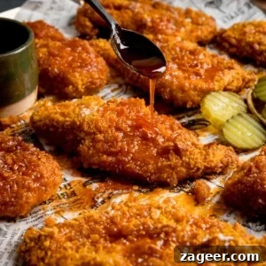 Spicy Honey-Glazed Chicken Recipe 8 Square image of crispy baked chicken breast fillets on a baking tray with sliced pickles. The chicken is being drizzled with hot honey sauce from a black spoon.