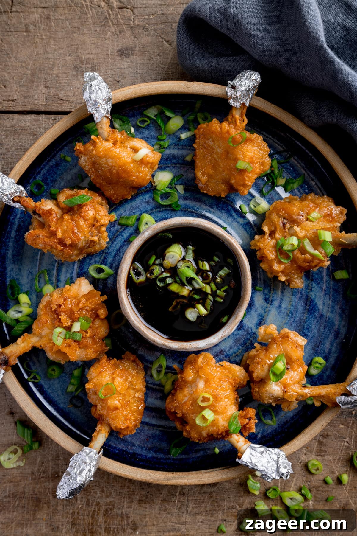 Crispy Chicken Lollipop Recipe: Irresistible Snack or Appetizer 5 Chicken lollipops arranged in a circle on a blue plate with a dipping bowl of sticky spicy sauce int he middle. The lollipops and sauce have been sprinkled with chopped spring onions and the plate is in a wooden table next to a blue napkin.