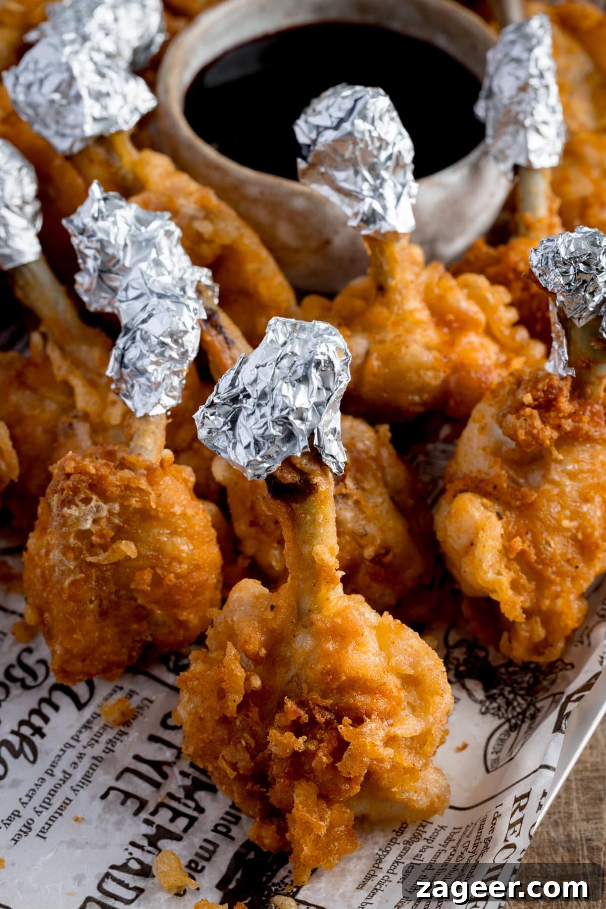 Crispy Chicken Lollipop Recipe: Irresistible Snack or Appetizer 6 Chicken lollipops, standing up on a piece of white baking paper that's decorated with newspaper-style writing. The lollipops have been wrapped in foil at the ends and they're next to a small light bowl of a dark dipping sauce.