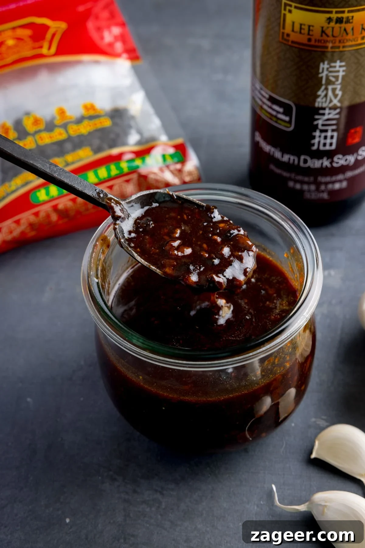 Authentic Black Bean Sauce Recipe for Stir-Fries and Marinades 2 A glass jar filled with homemade black bean sauce. There is a black spoon, taking a spoonful from the jar.
The jar is on a dark surface and there are ingredients scattered around.