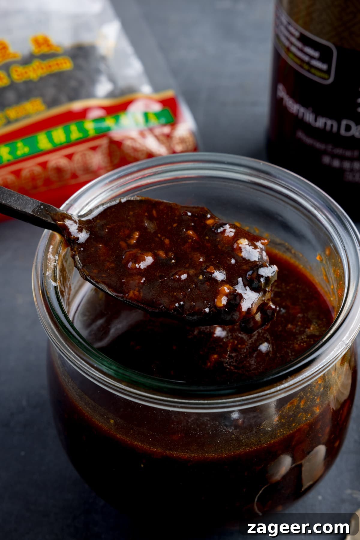 Authentic Black Bean Sauce Recipe for Stir-Fries and Marinades 6 A glass jar filled with homemade black bean sauce. There is a black spoon, taking a spoonful from the jar. The jar is on a dark surface and there are ingredients in the background.