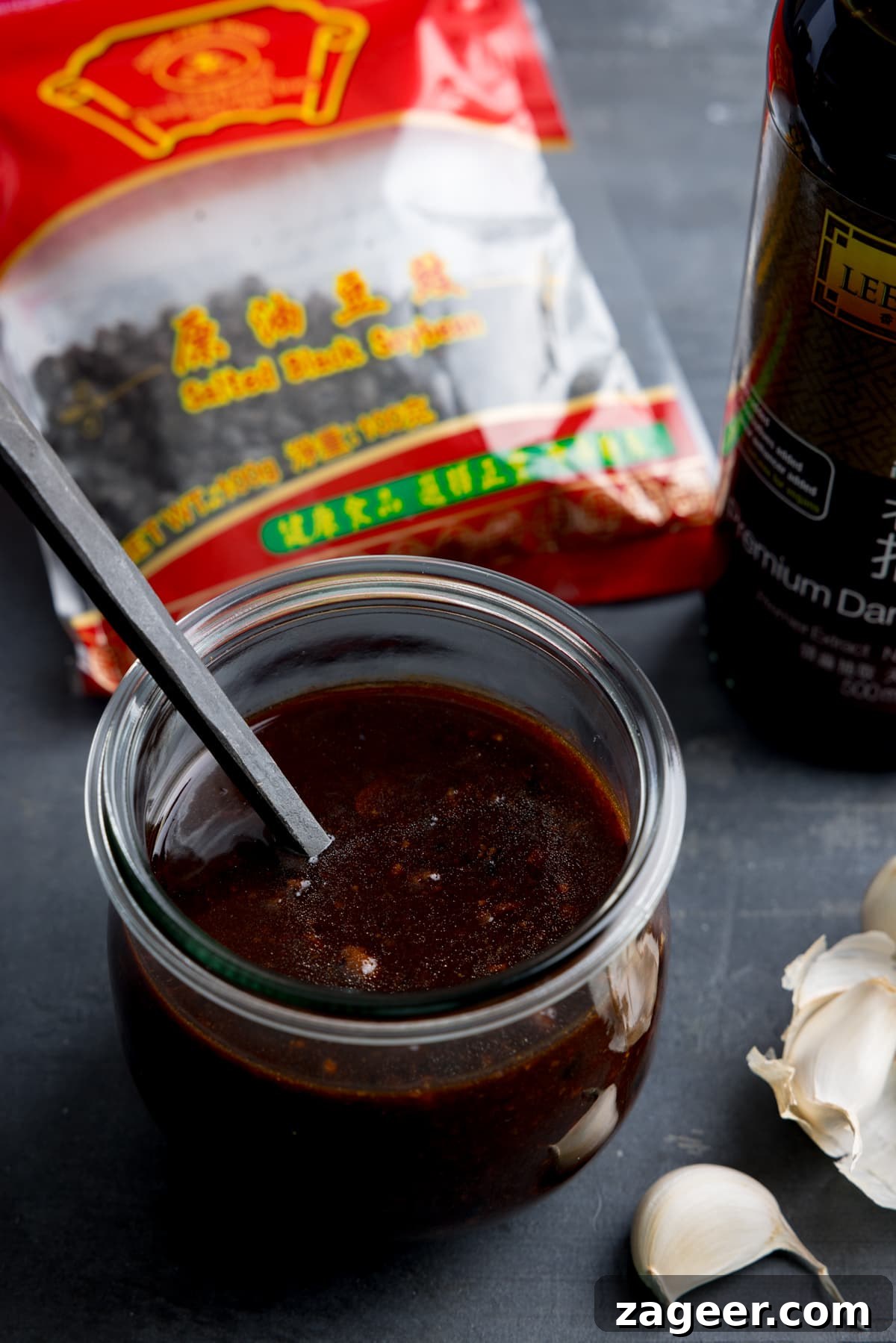 Authentic Black Bean Sauce Recipe for Stir-Fries and Marinades 8 A glass jar filled with homemade black bean sauce. There is a black spoon sticking out of the jar. The jar is on a dark surface and there are ingredients in the background