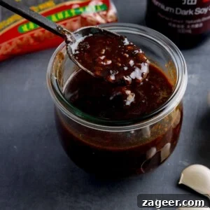 Authentic Black Bean Sauce Recipe for Stir-Fries and Marinades 9 square image of a glass jar filled with homemade black bean sauce. There is a black spoon, taking a spoonful from the jar. The jar is on a dark surface and there are ingredients in the background