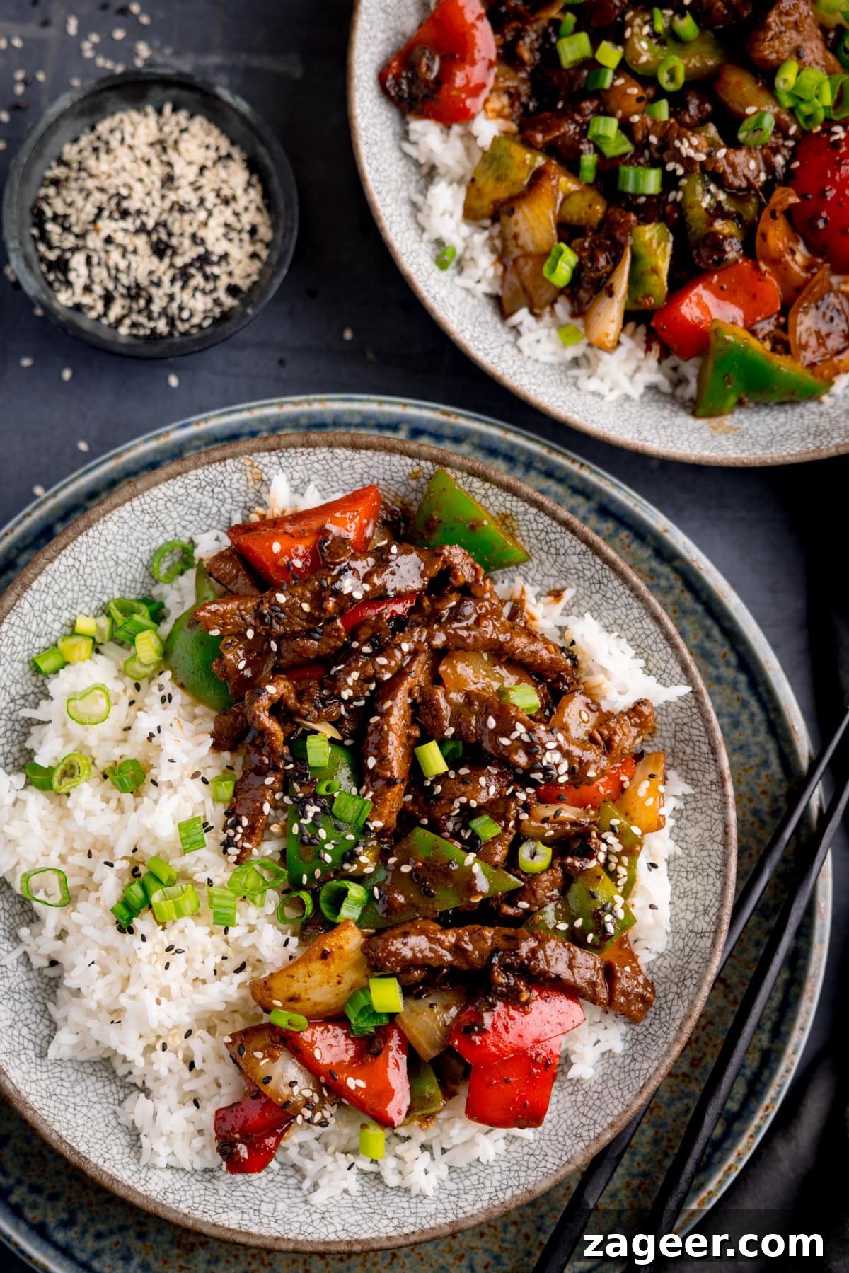 Stir-Fried Beef in Savory Black Bean Sauce Recipe 2 Beef in black bean sauce stir fry with boiled rice in a bowl, topped with spring onions and sesame seeds. The bowls is on a dark surface and there is a further bowl in shot. There is a small bowl of black and white sesame seeds nearby.