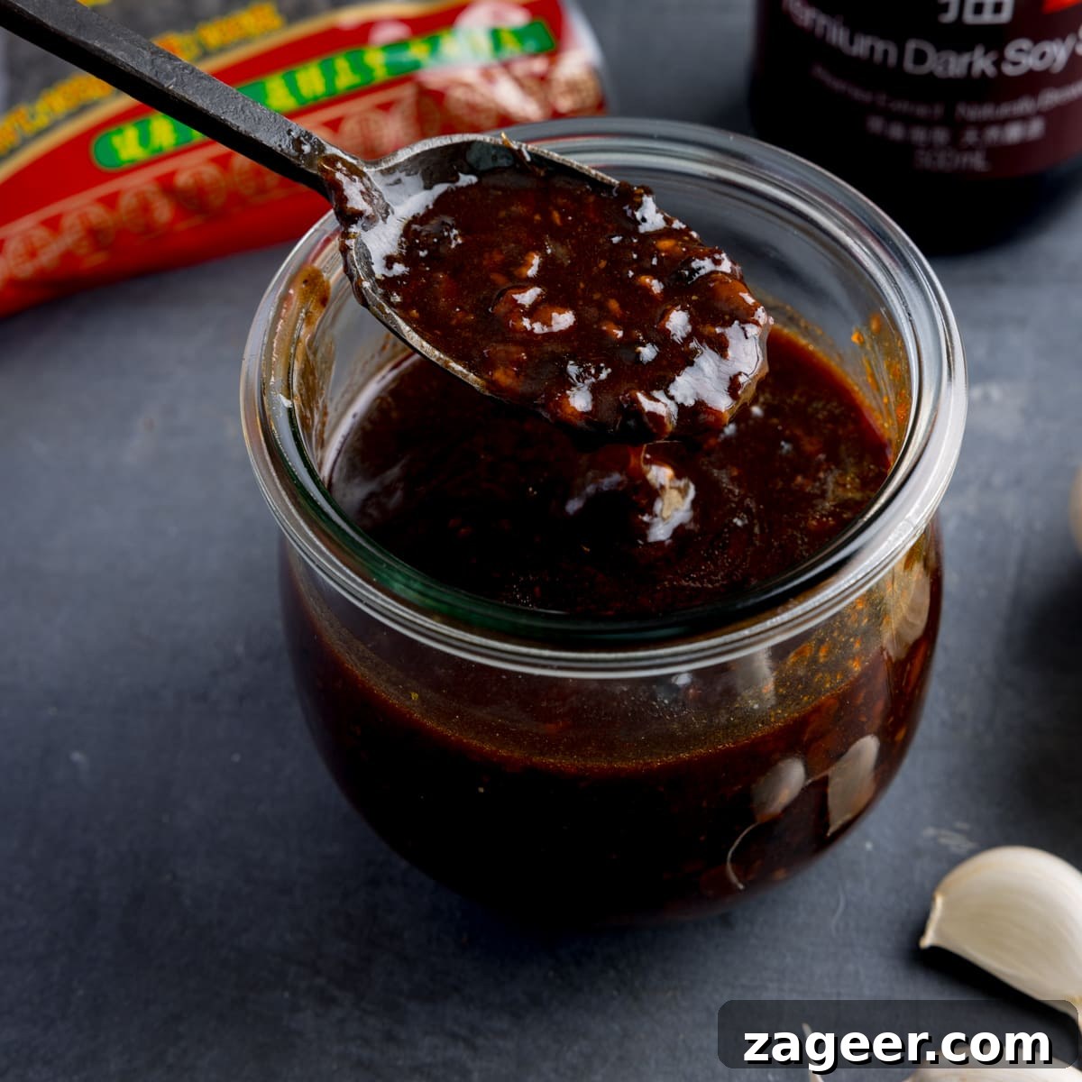 Stir-Fried Beef in Savory Black Bean Sauce Recipe 5 square image of a glass jar filled with homemade black bean sauce. There is a black spoon, taking a spoonful from the jar. The jar is on a dark surface and there are ingredients in the background